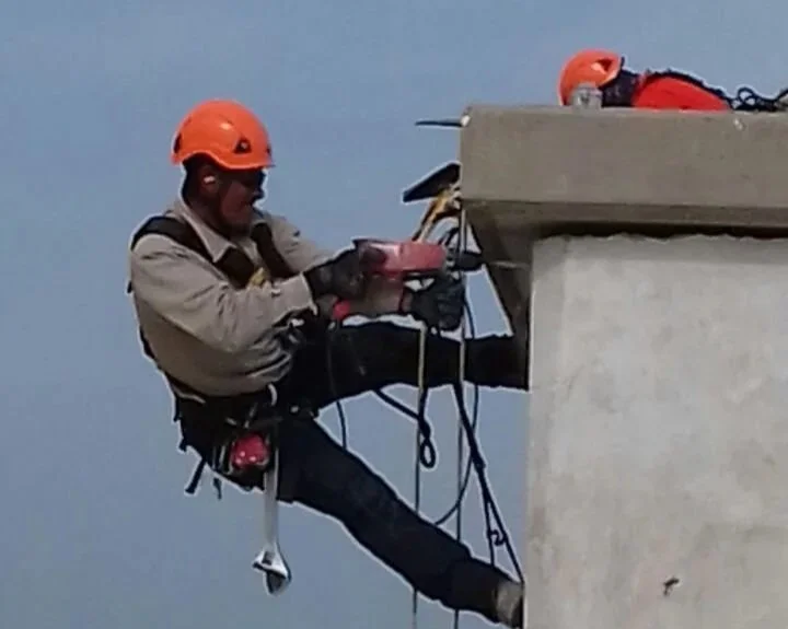 Trabajador en altura con casco naranja, usando arnés y equipo de protección, inspecciona o trabaja en la estructura de un edificio.