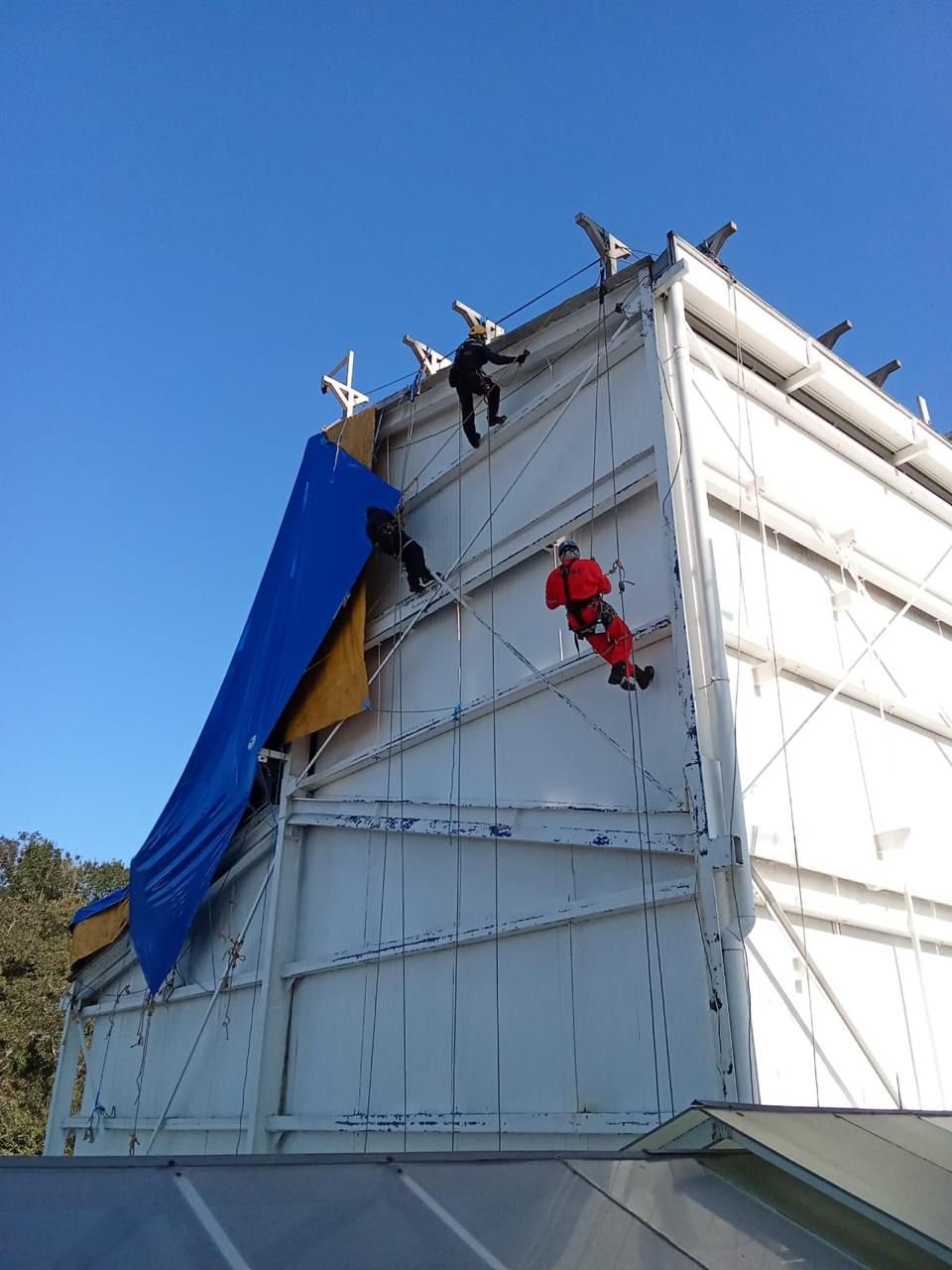 Tres personas trabajando en altura en la estructura de un edificio, usando arneses y cuerdas de seguridad.