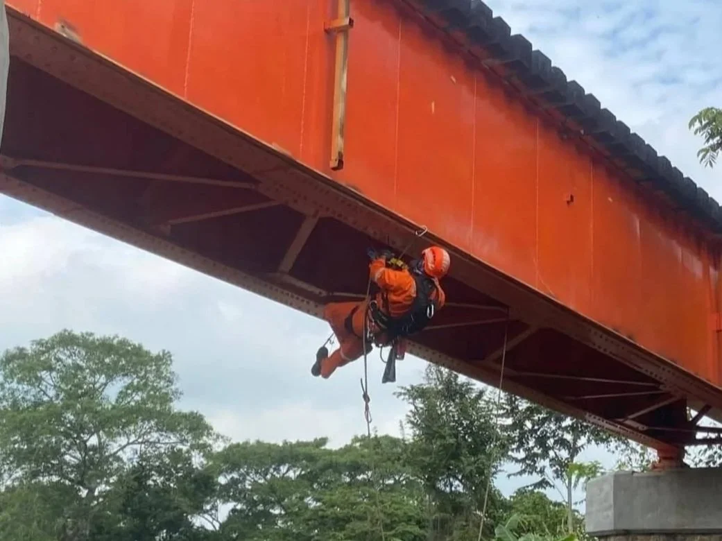 Trabajador en arnés inspeccionando la estructura de un puente de hierro rojo desde abajo.
