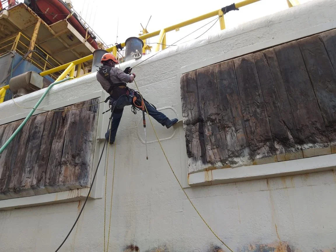 Un trabajador de reparación en una embarcación, usando equipo de seguridad, sostiene una cuerda y realiza tareas de mantenimiento en la estructura del barco.