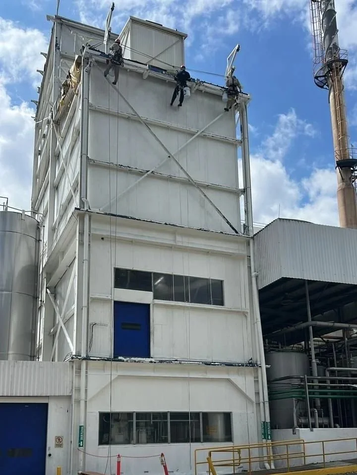 Varios trabajadores en el techo de una estructura industrial, realizando tareas de mantenimiento o reparación en una planta industrial con cielo despejado.