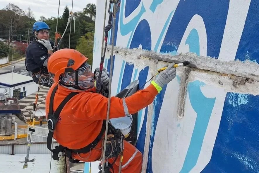 Dos trabajadores realizando labores de mantenimiento en una pared exterior, uno pintando y otro observando, con equipo de protección y en una estructura elevada.
