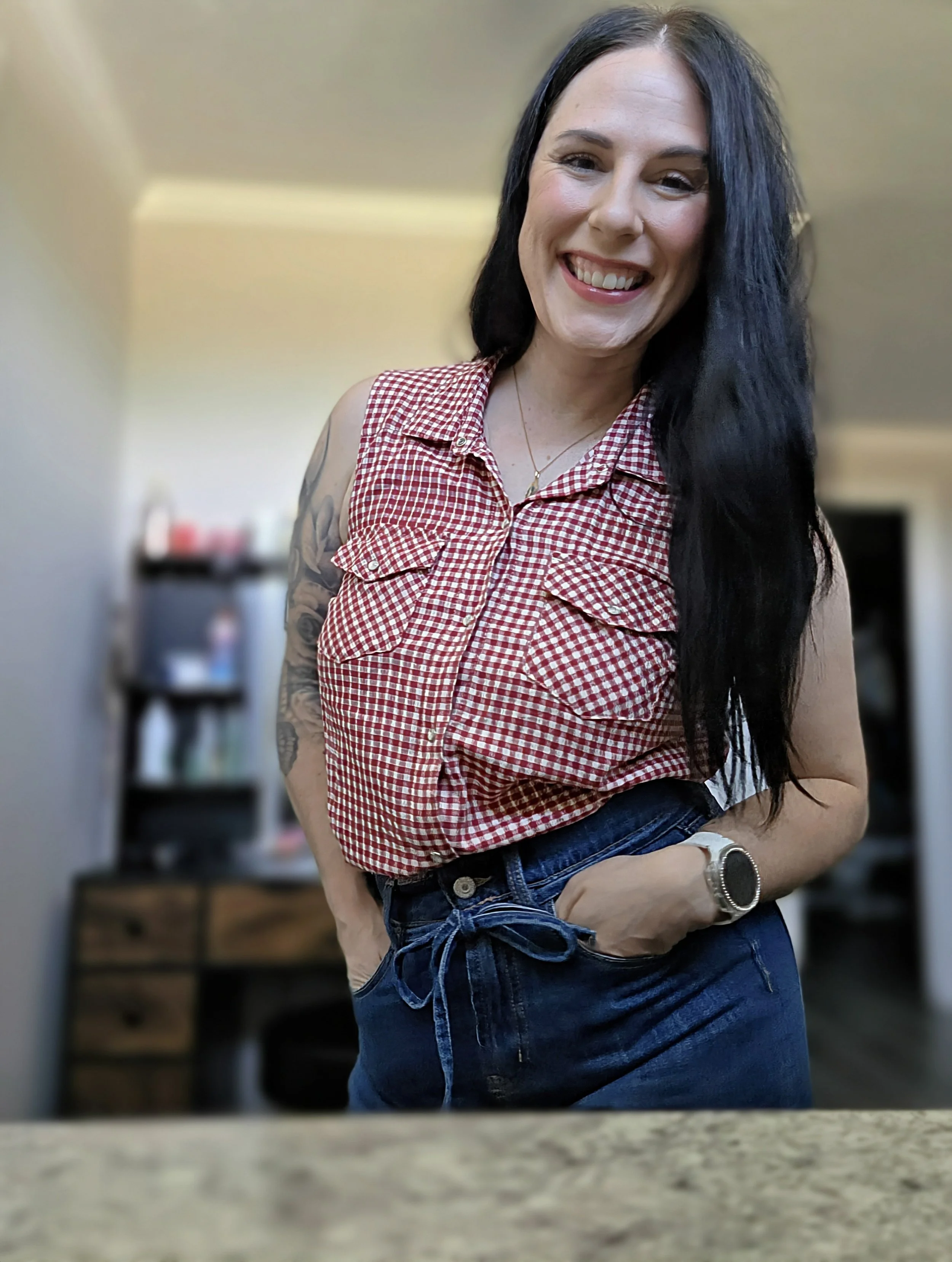 A woman with long black hair smiling, wearing a red and white checkered sleeveless top and blue jeans, standing in a room with blurred background including a bookshelf and television.