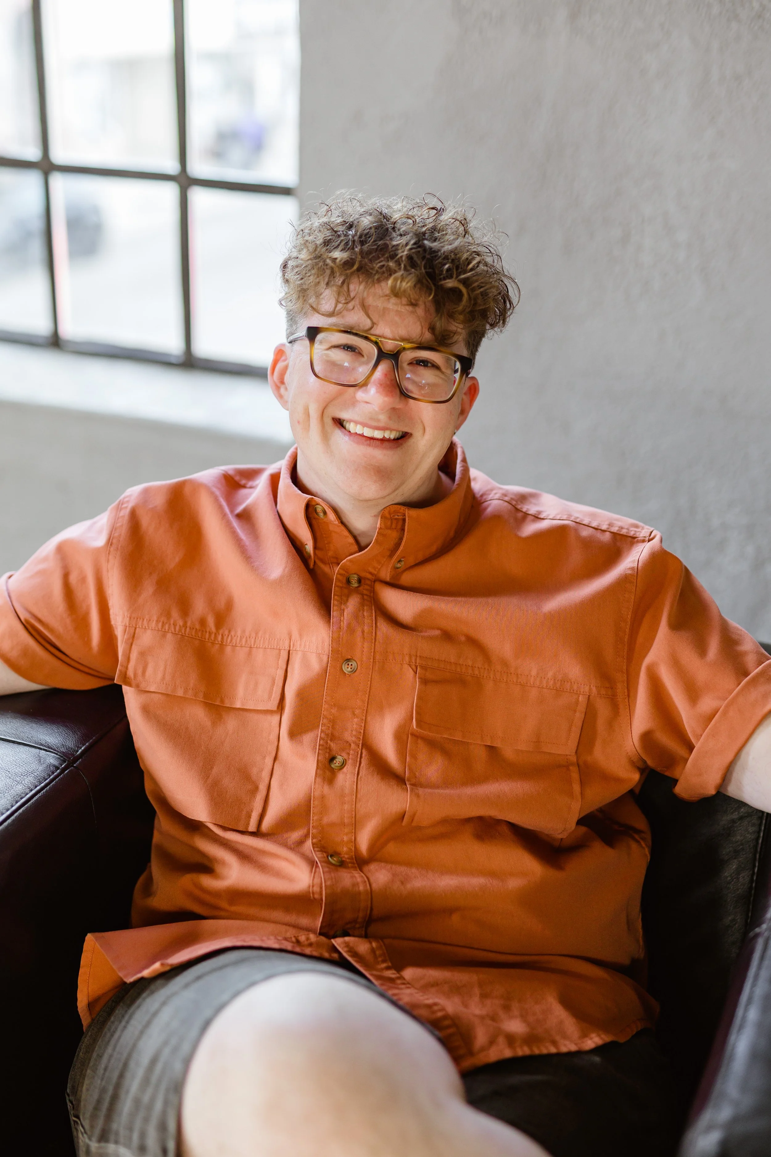 A smiling young man with curly hair and glasses, wearing an orange shirt, sitting on a black sofa inside a room with a large window.