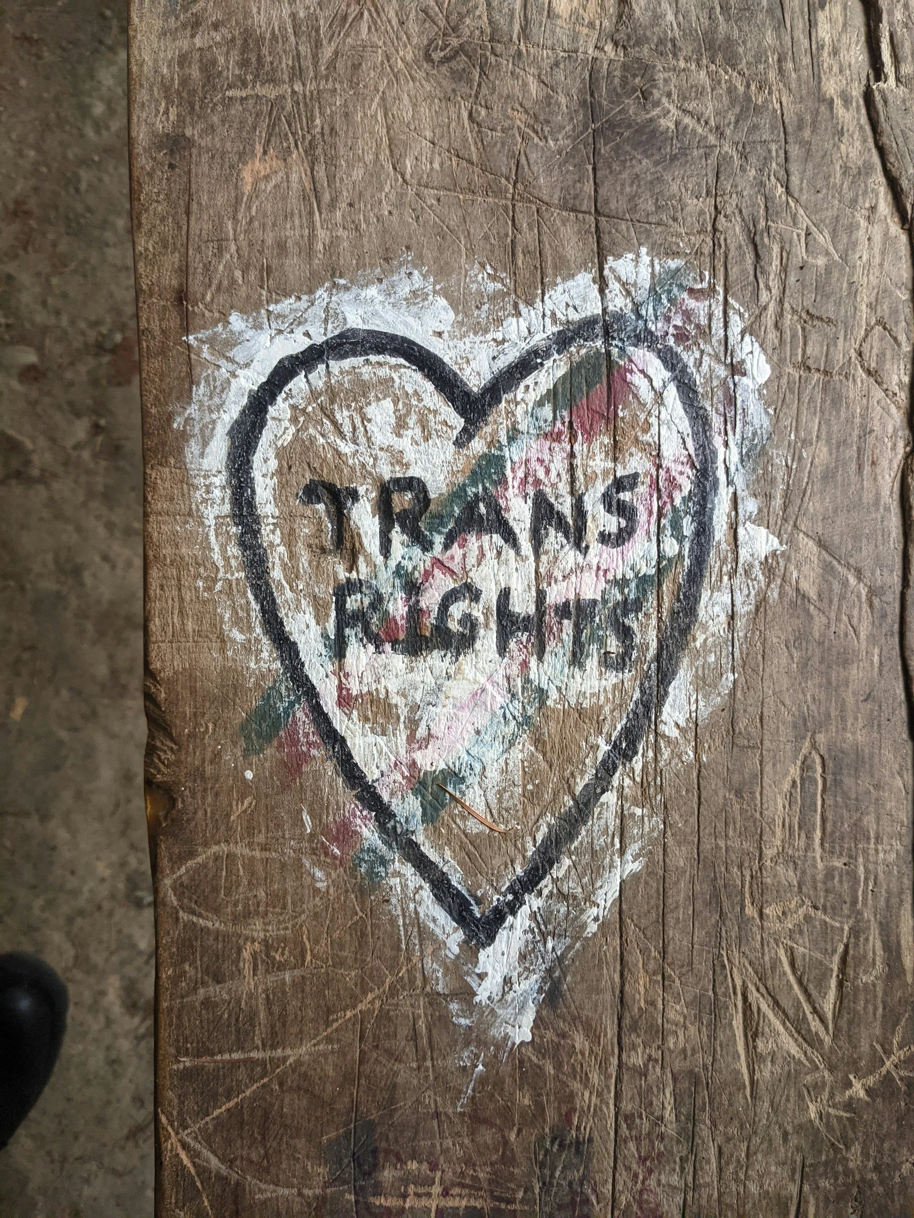 A piece of wood with graffiti that says 'Trans Rights' inside a heart, along with some white paint splatters and markings.