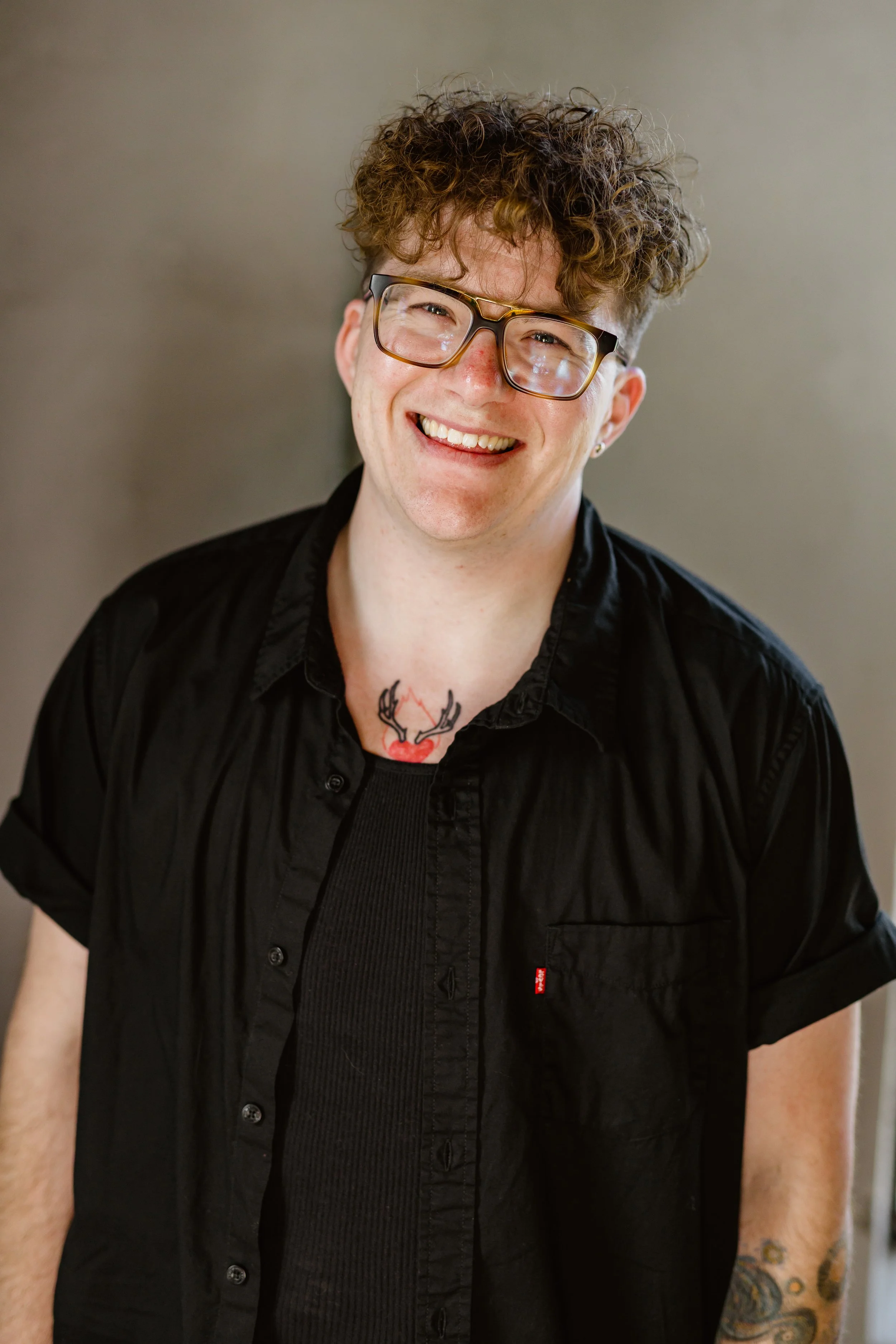 A young man with curly brown hair, glasses, and a big smile, wearing a black shirt and showing tattoos on his chest and arm.