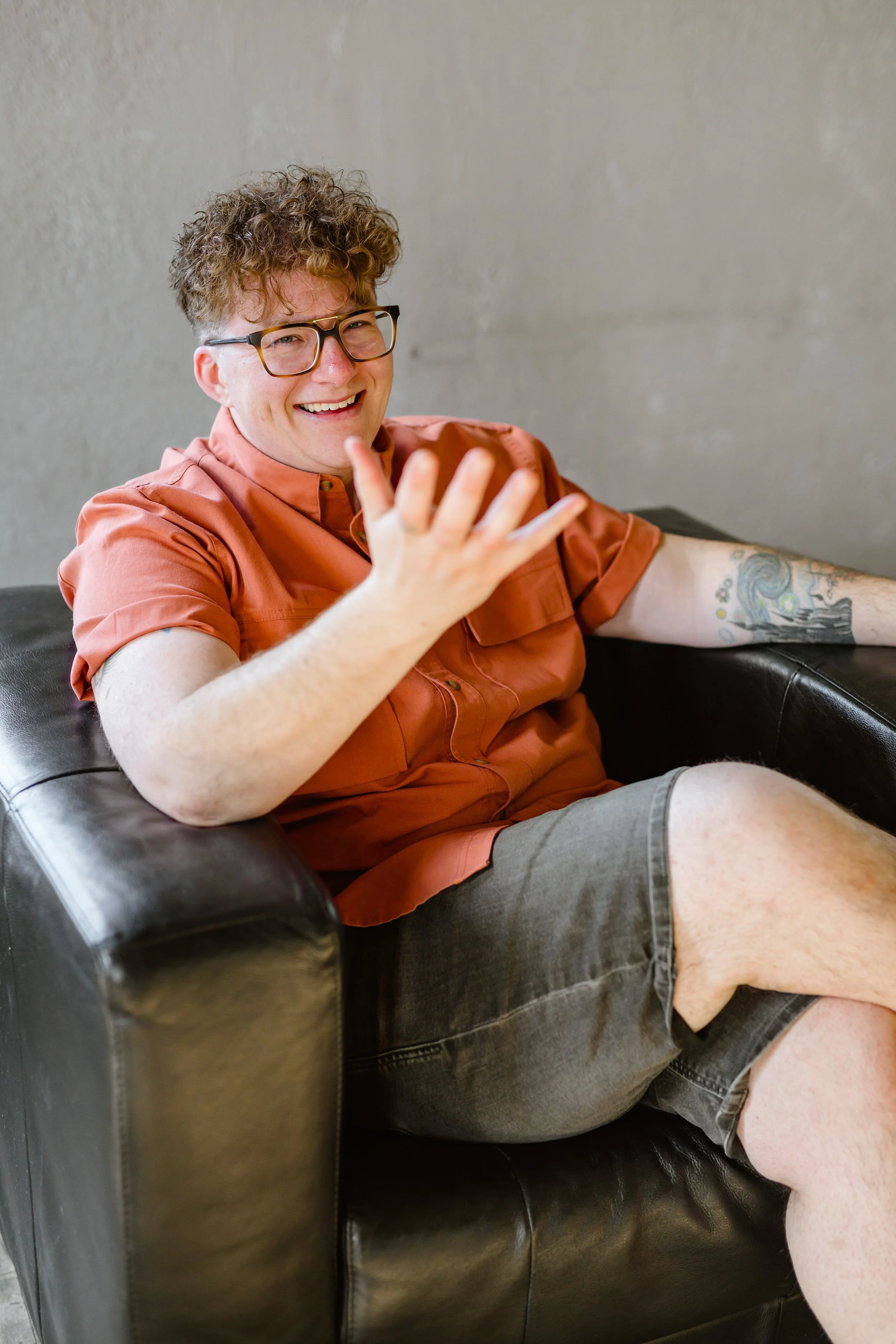 A young man with curly hair, glasses, and a big smile, sitting on a black leather sofa, wearing a short-sleeved orange shirt and gray shorts, gesturing with his right hand, with tattoos on his left arm, against a plain beige wall.