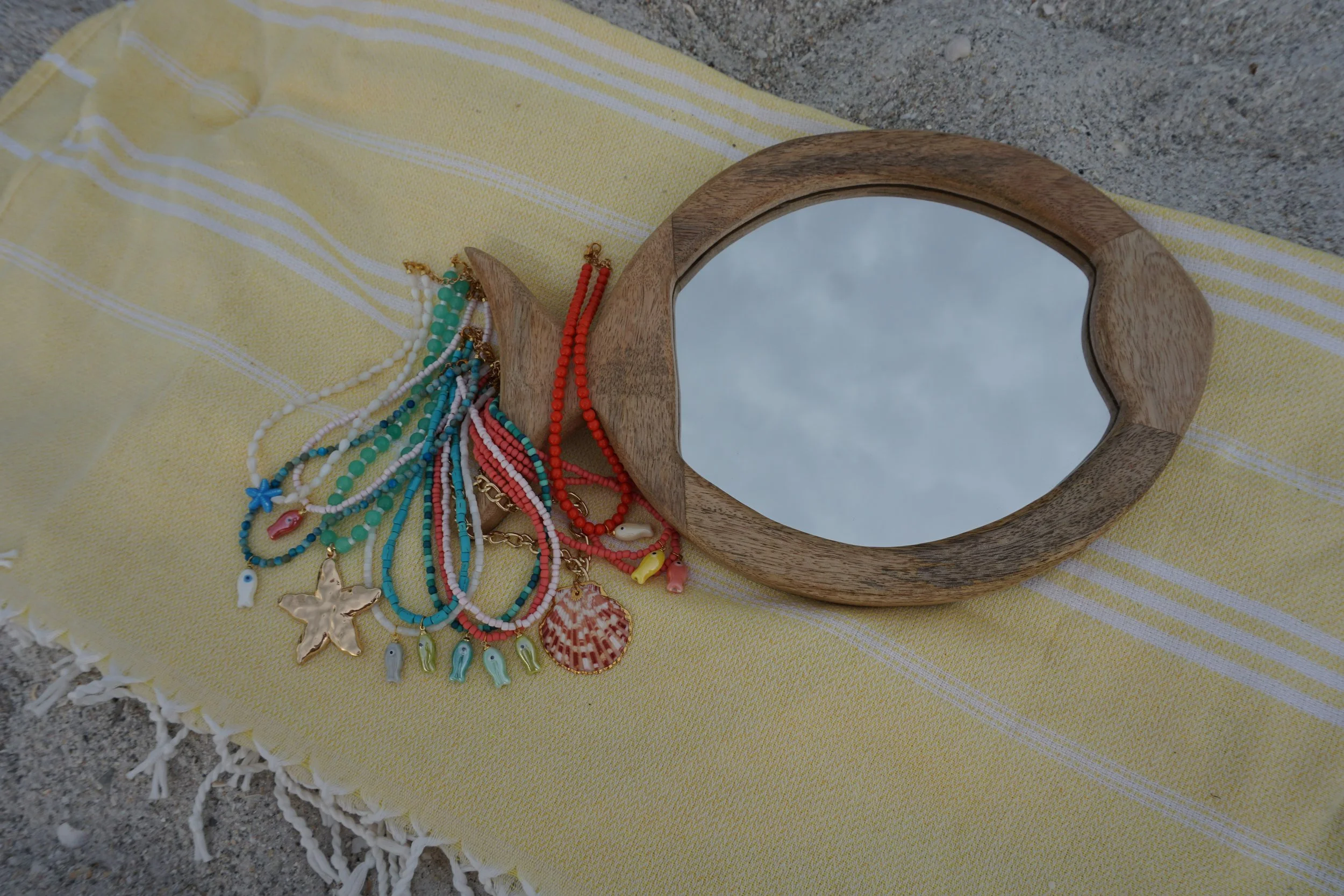 Colorful beaded necklaces with seashell charms spread on a yellow and white striped cloth, with a wooden mirror reflecting the sky and clouds, on a sandy surface.