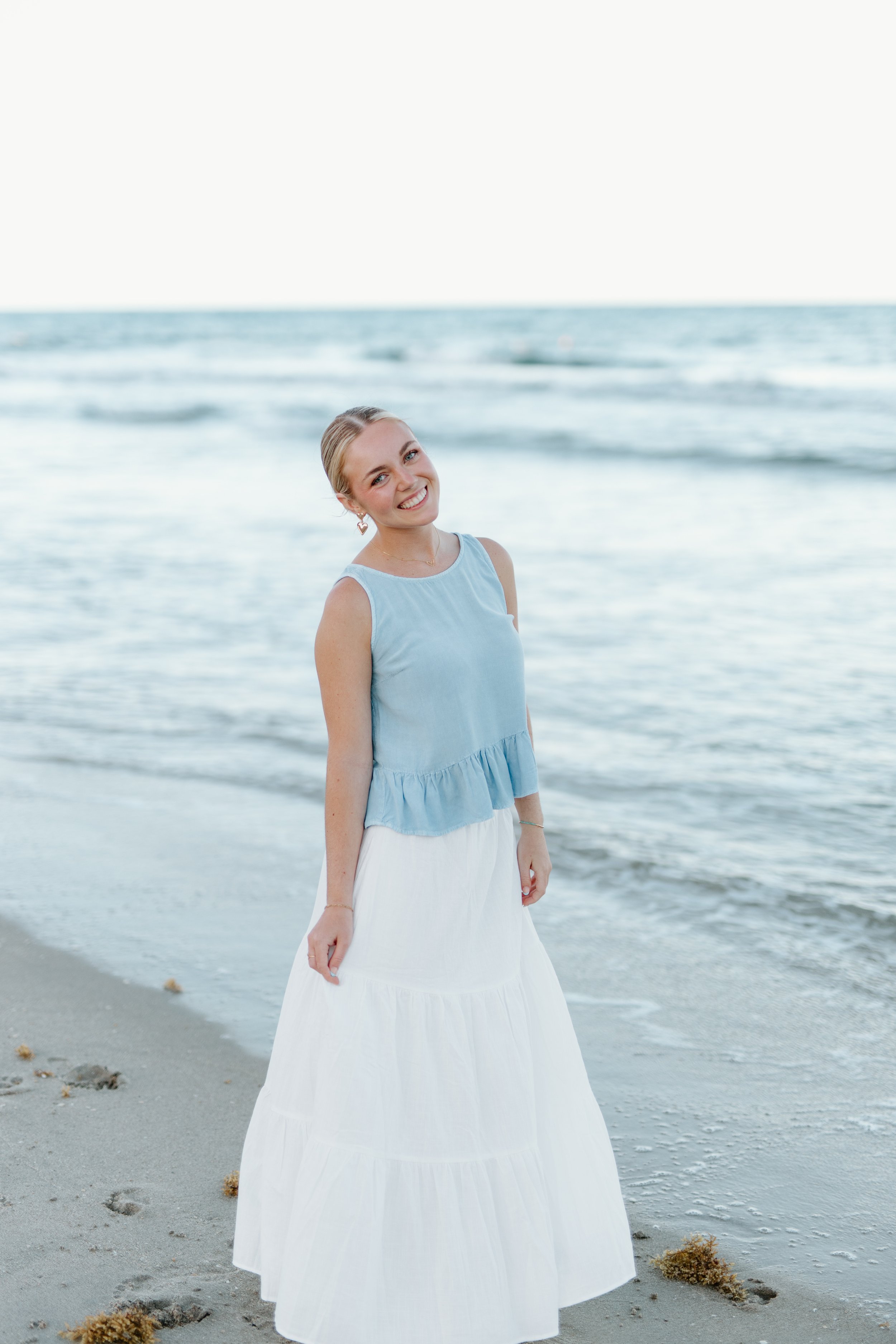 A young woman with blonde hair, wearing a sleeveless blue top and a long white skirt, stands on a sandy beach near the ocean, smiling at the camera.