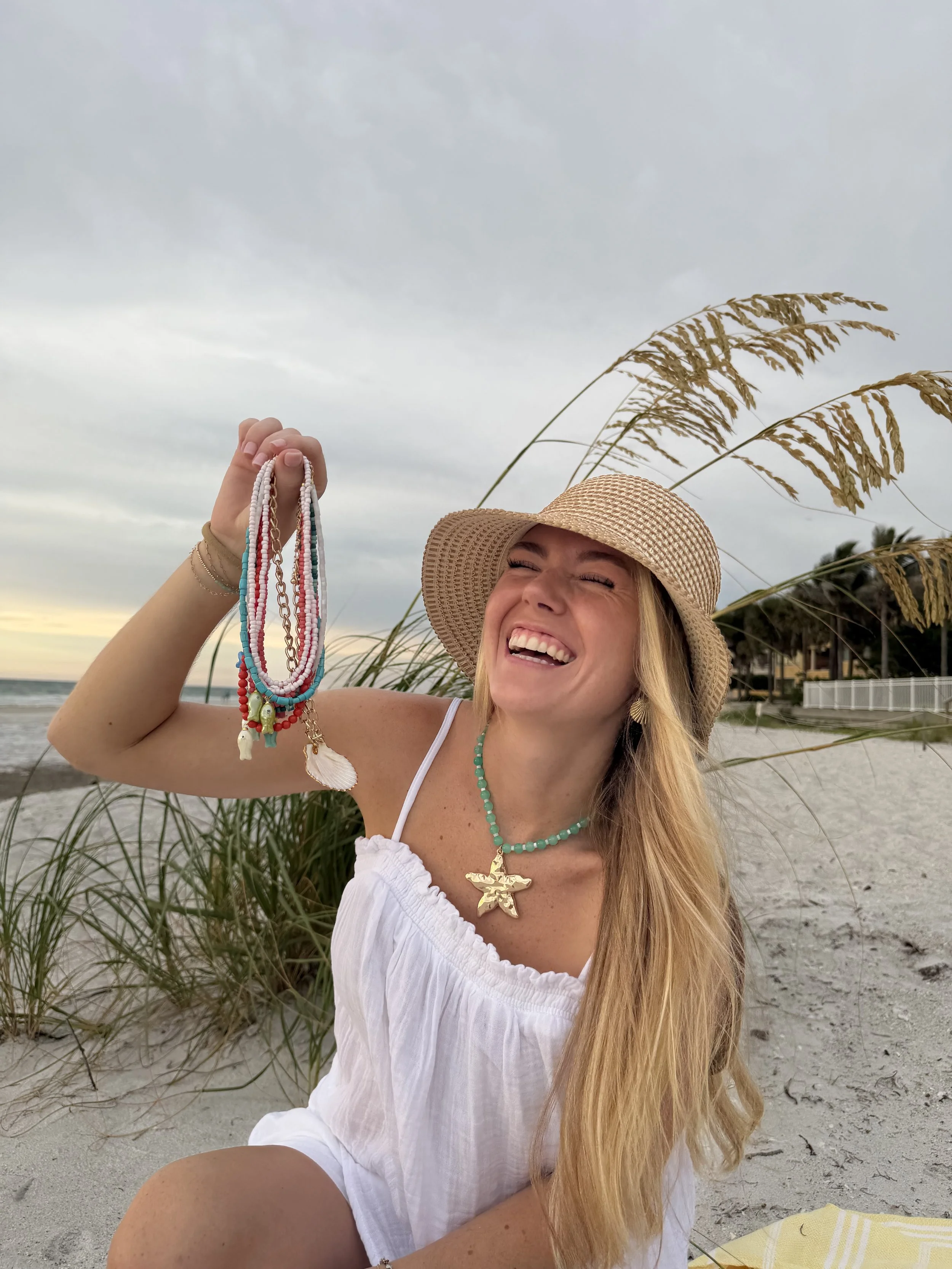 A woman with long blonde hair smiling and laughing at the beach, wearing a straw hat, white dress, turquoise necklace with a starfish pendant, and holding colorful beaded necklaces, with sand, grass, and cloudy sky in the background.