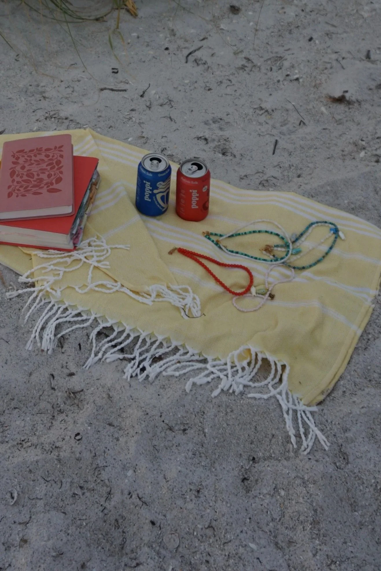 A yellow towel with white stripes and fringe edges is spread on the sandy ground, holding two soda cans (one blue Poppi and one red Poppi) at the beach. Also on the towel are the Bible, and beaded necklaces.
