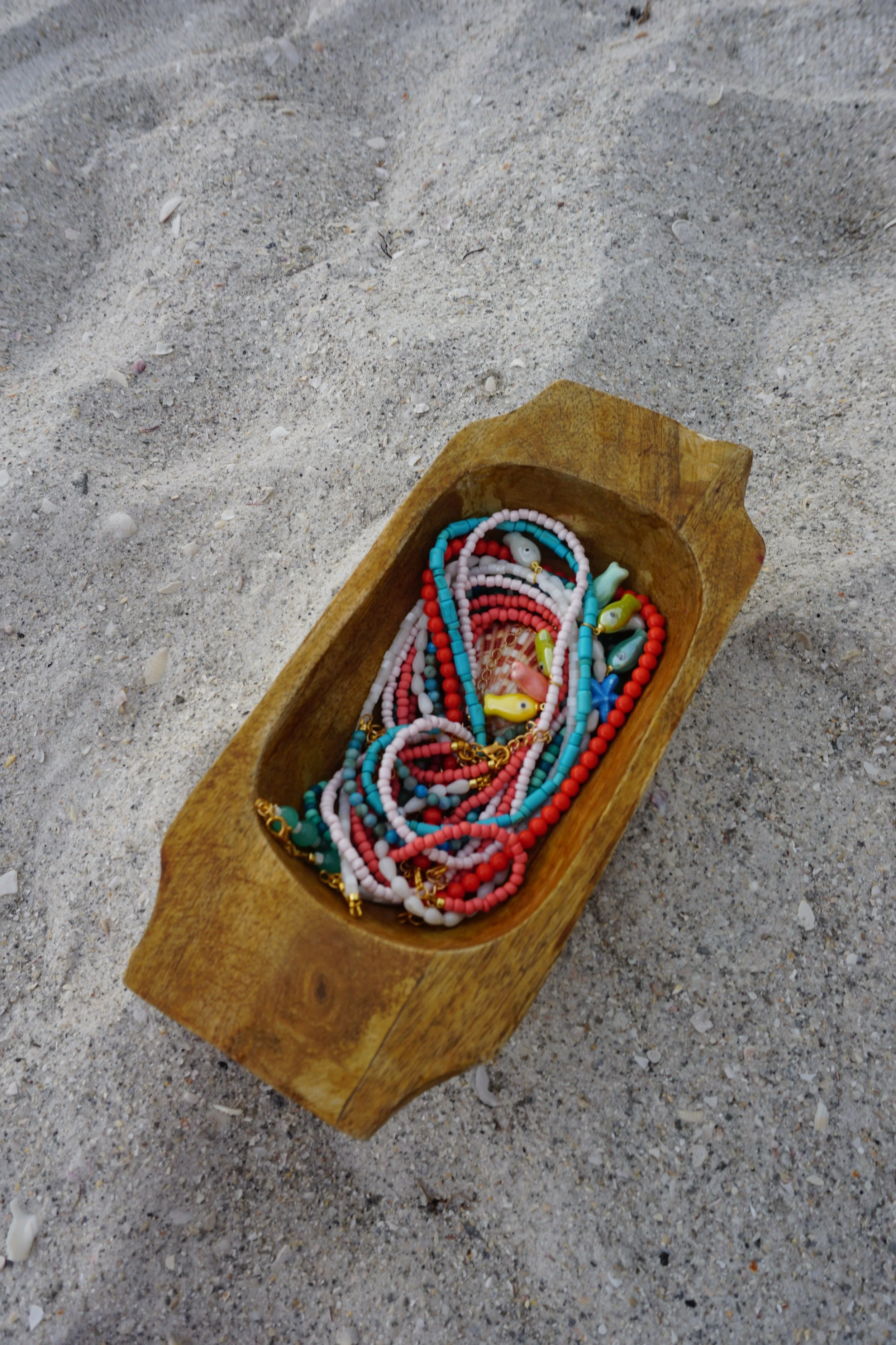 Colorful beaded necklaces with fish-shaped pendants inside a wooden container on sandy beach.