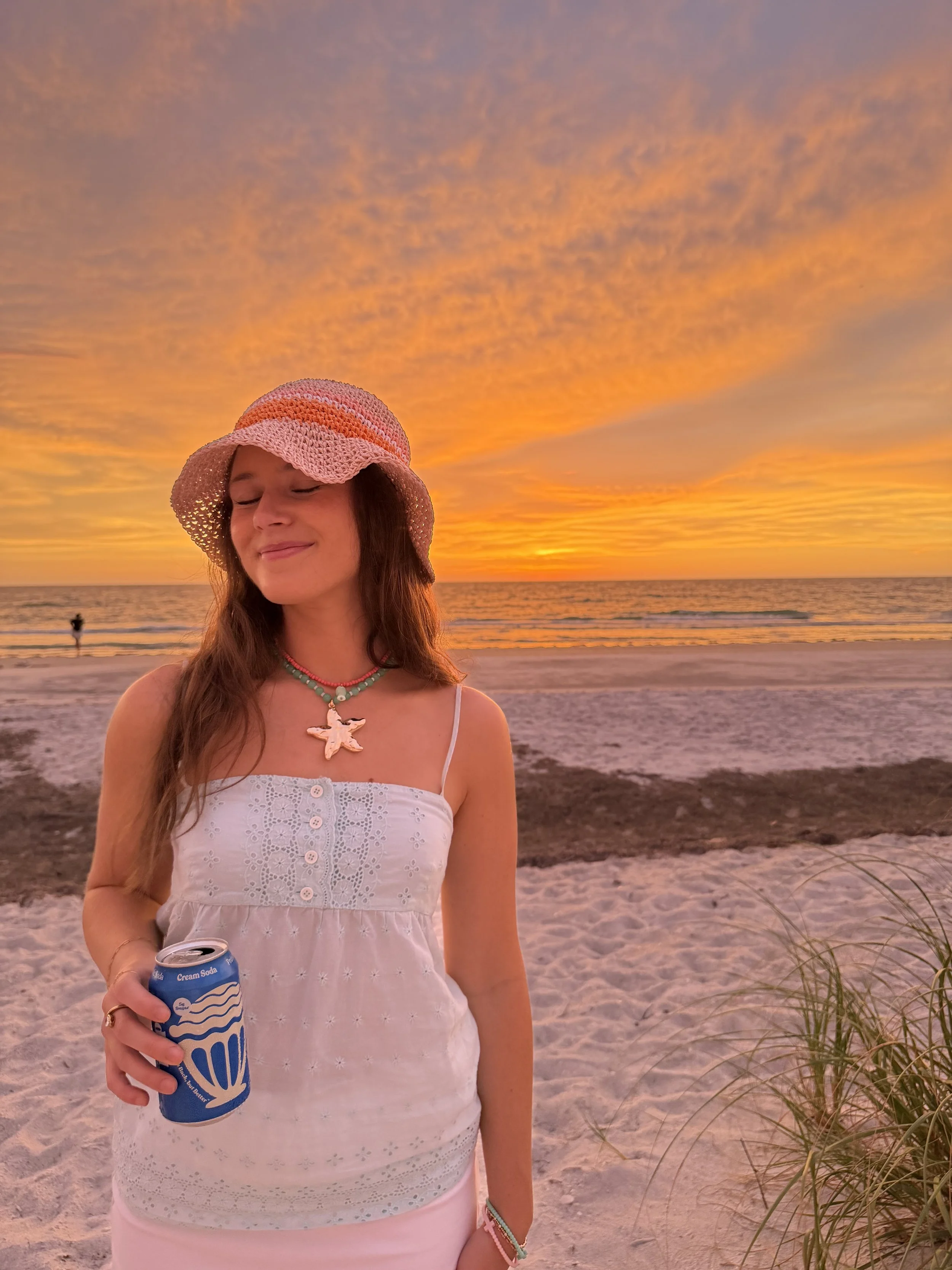 A young woman with long brown hair wearing a pink and orange knitted sunhat, a light-colored dress, and colorful beaded necklaces, standing on the beach at sunset, holding a can of cream soda, with the ocean and colorful sky in the background.
