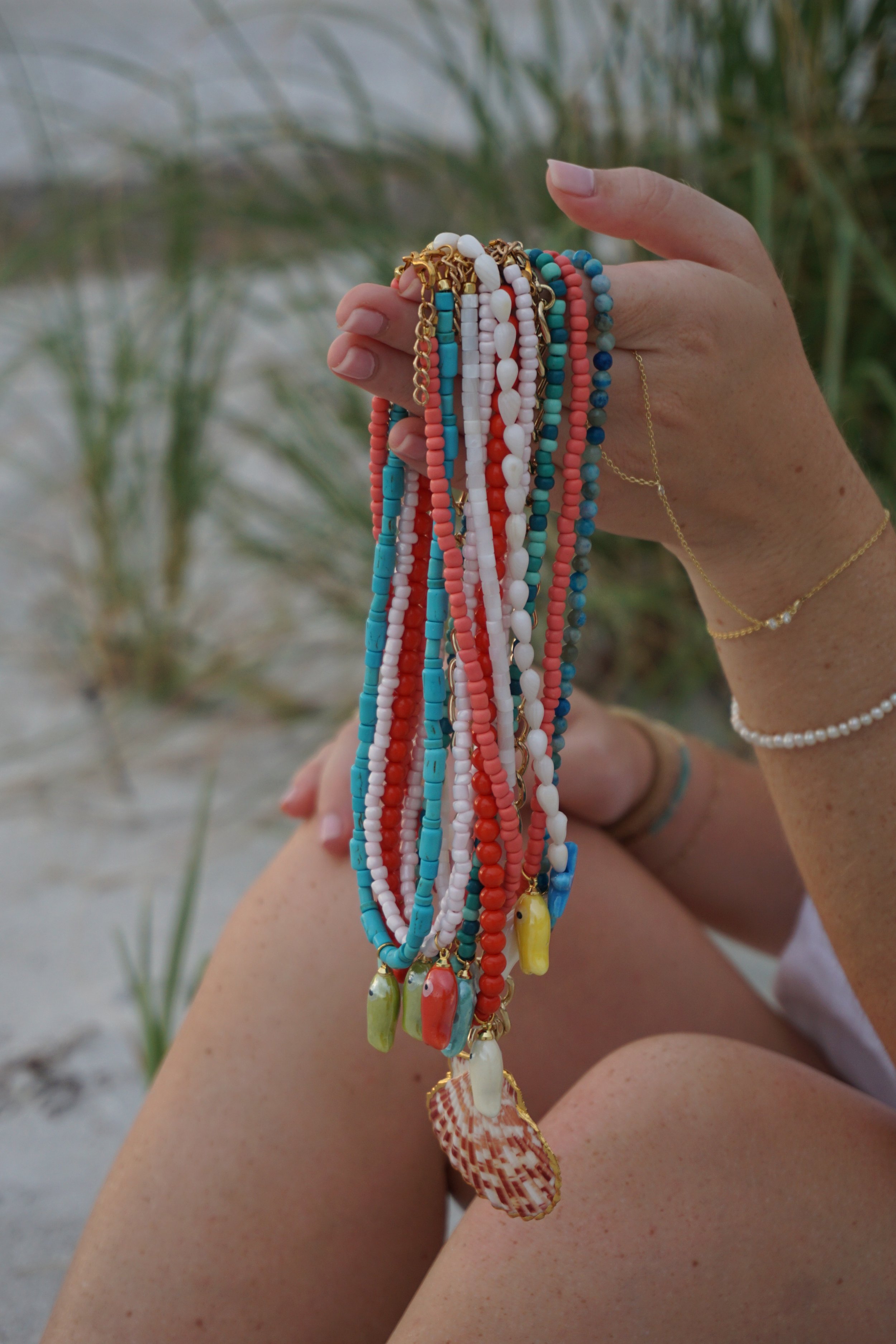 Person holding colorful beaded necklaces outdoors near sand and grass, with visible arm and part of leg, and a gold bracelet and ring.