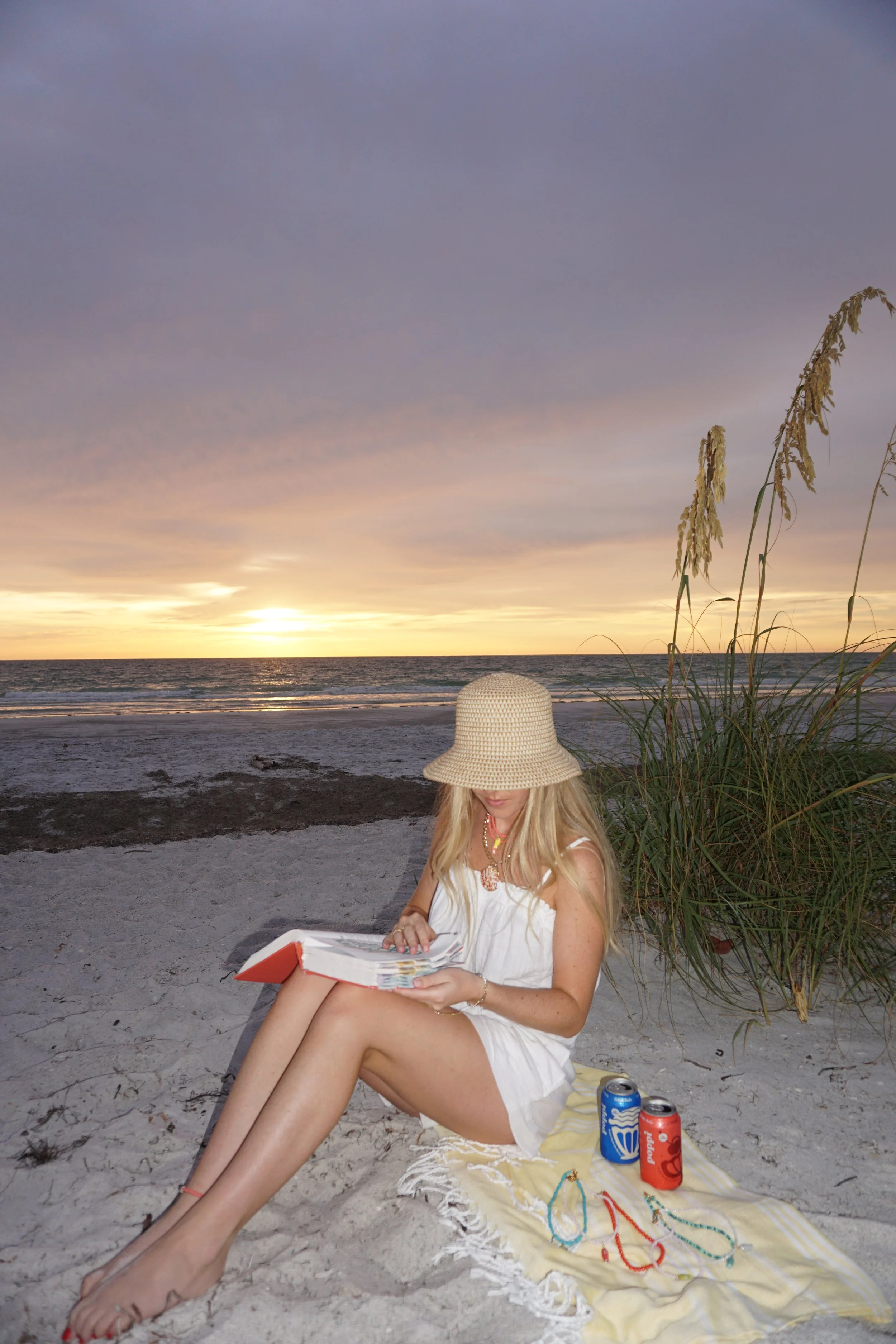 A woman in a white dress and straw hat sitting on a towel on the beach, reading the Bible, with two cans of beverages beside her, near the ocean at sunset.