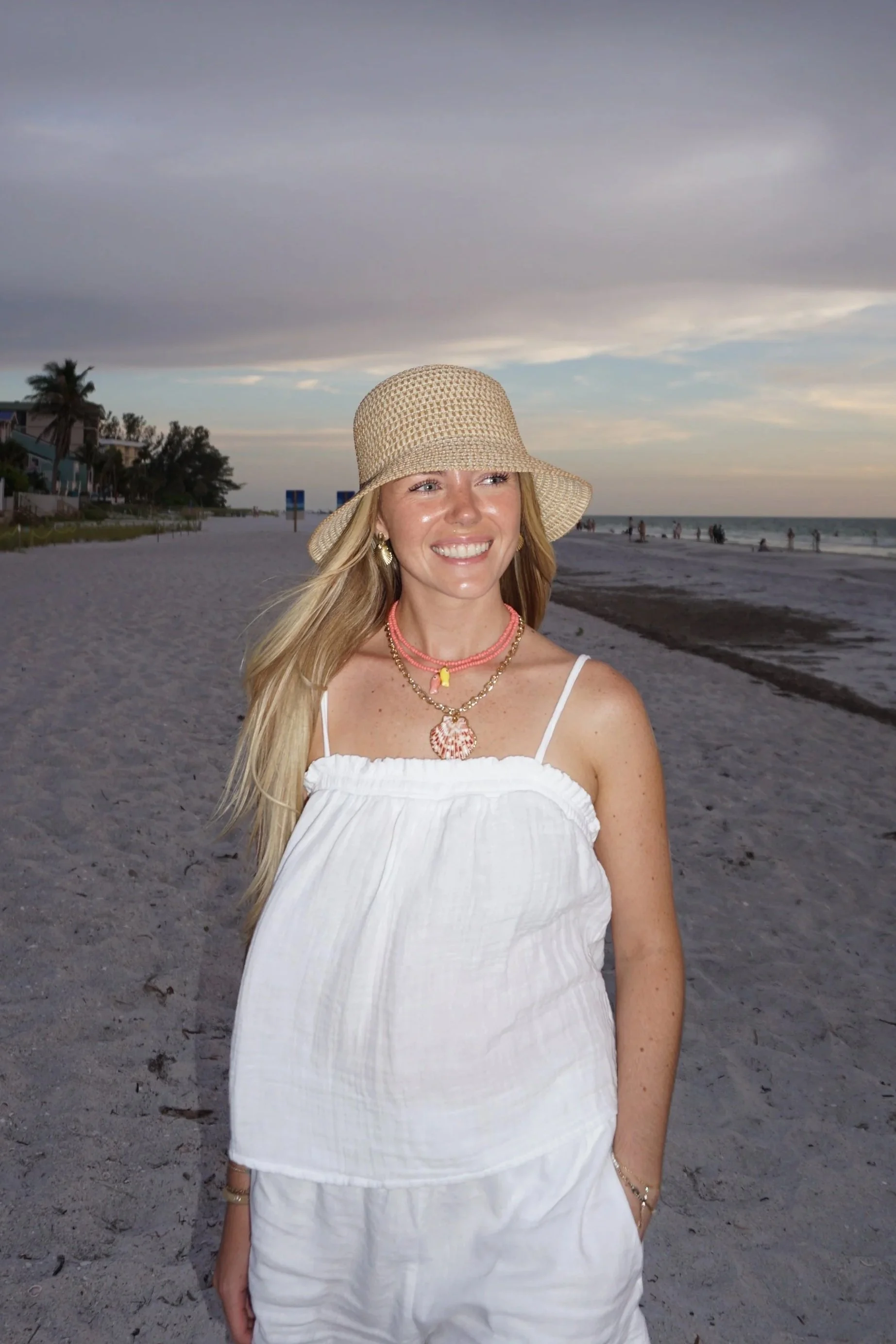 A woman smiling on a beach with a straw sun hat, white dress, and layered necklaces, with houses, palm trees, and waves in the background during a cloudy day with fading light.