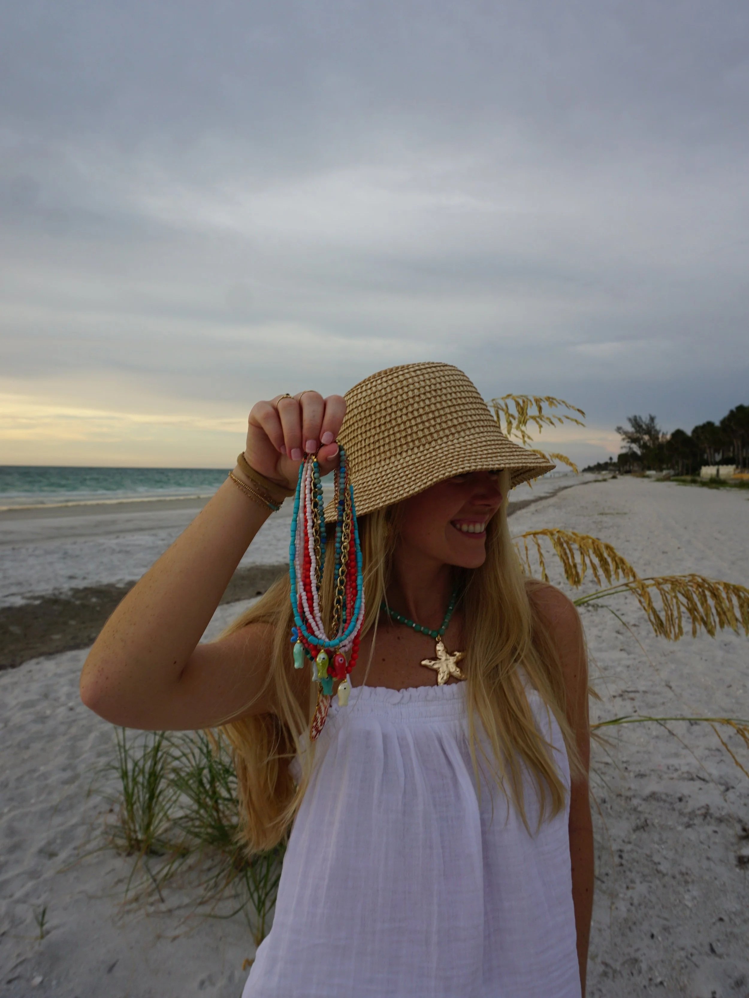 A woman on a beach holding colorful beaded jewelry and wearing a wide-brimmed hat, with the ocean and cloudy sky in the background.