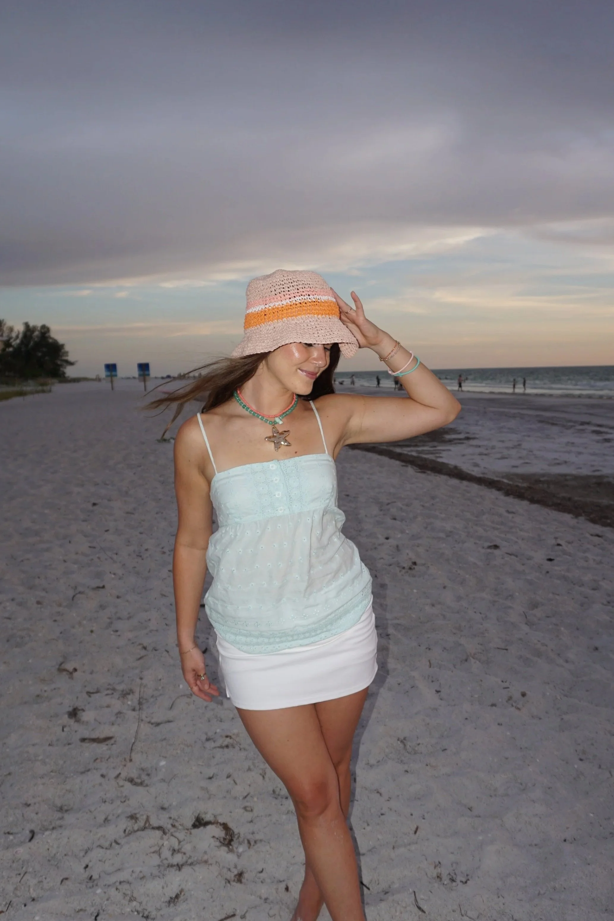 A woman at the beach wearing a sunhat, white dress, and fun jewelry, with the ocean, sky, and clouds in the background.