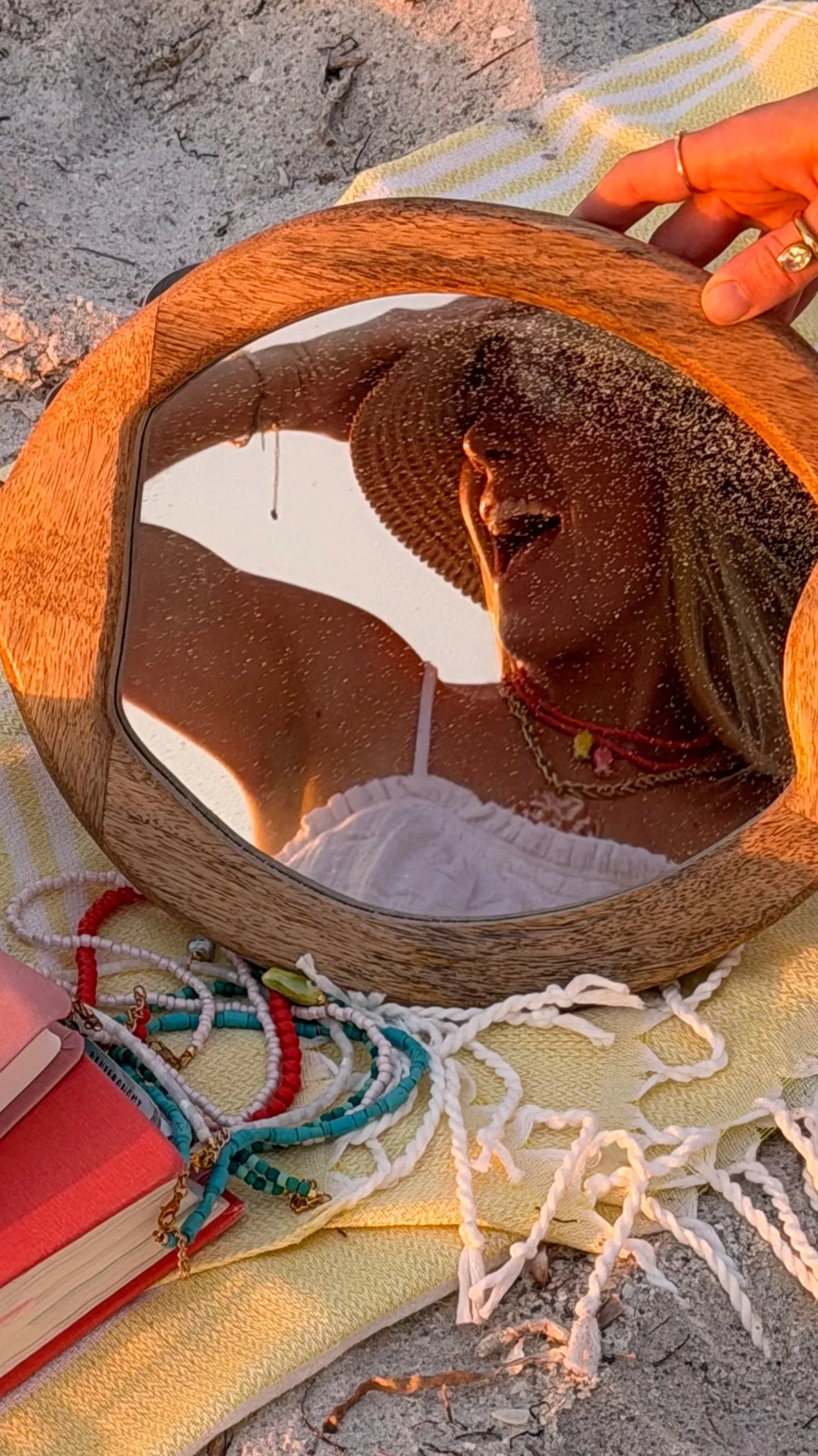 A mirror reflecting two people wearing a woven hat, surrounded by colorful beaded jewelry, the Bible, and a towel, placed on the sand.
