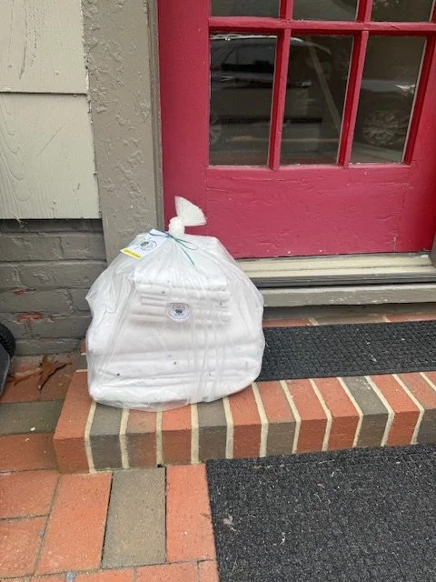 A large clear plastic trash bag with folded sheets inside placed on the concrete step outside a building with a red door and brick foundation.