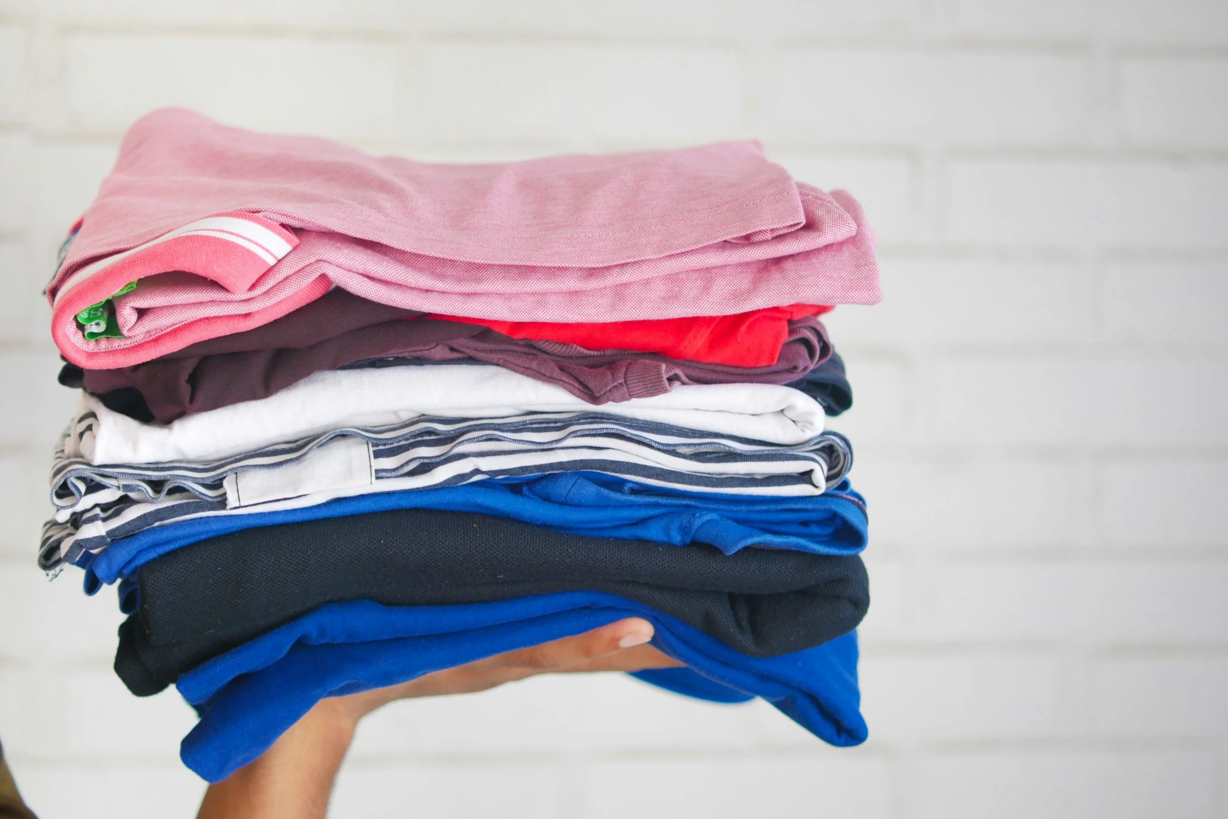 A person holding a neatly folded stack of various colored clothes against a white background.