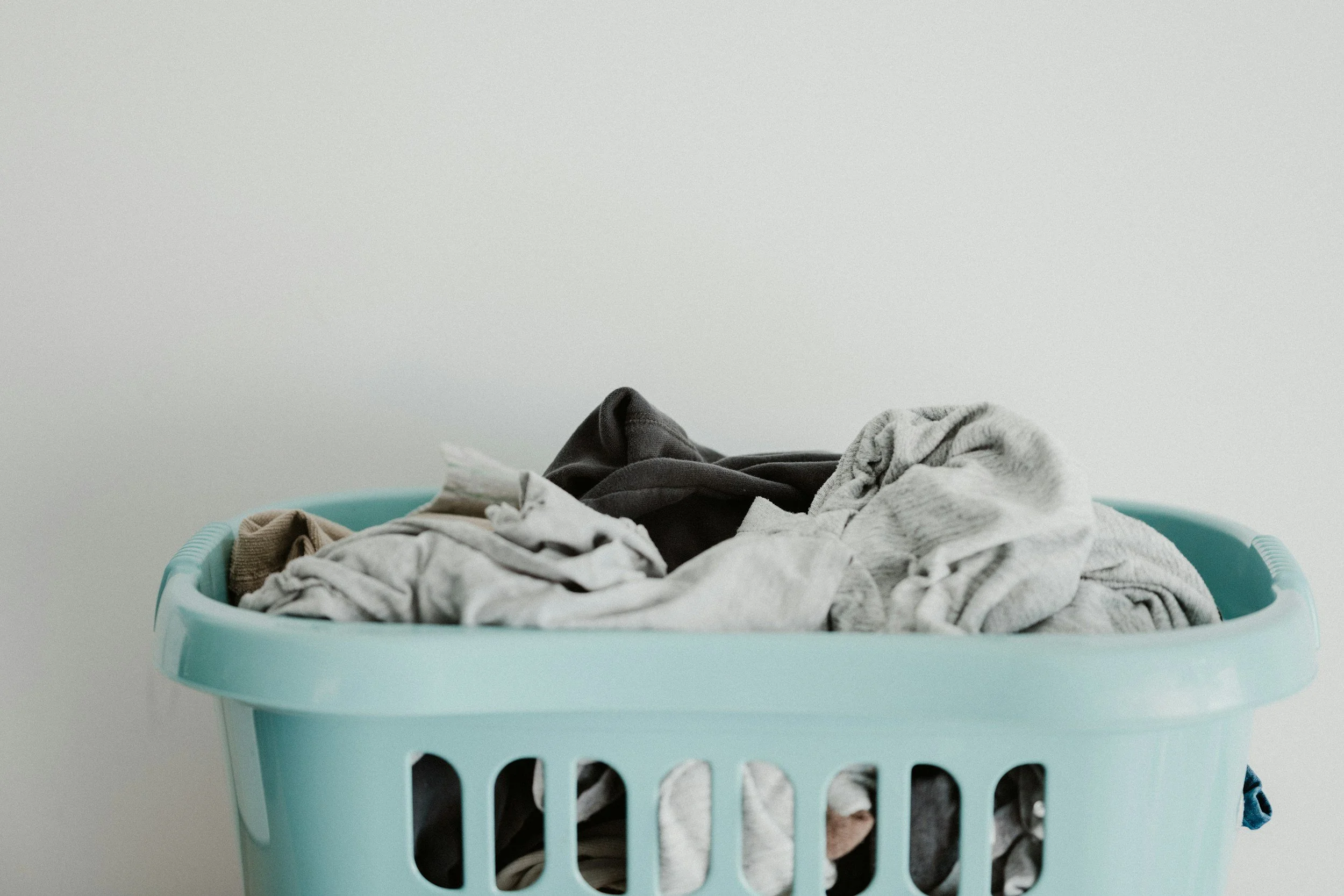 A laundry basket filled with folded clothes and laundry, with some hanging out over the edge. The basket is turquoise, and the background is a plain white wall.