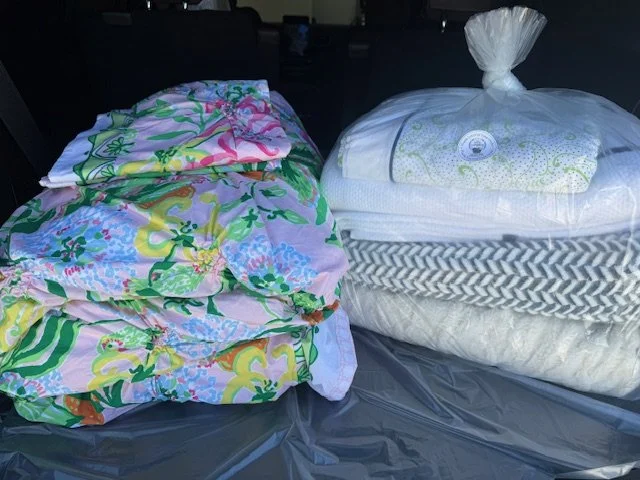 Bundles of household items, including folded bed linens with colorful floral patterns on the left and wrapped bedding and pillows on the right, placed on a dark plastic surface.