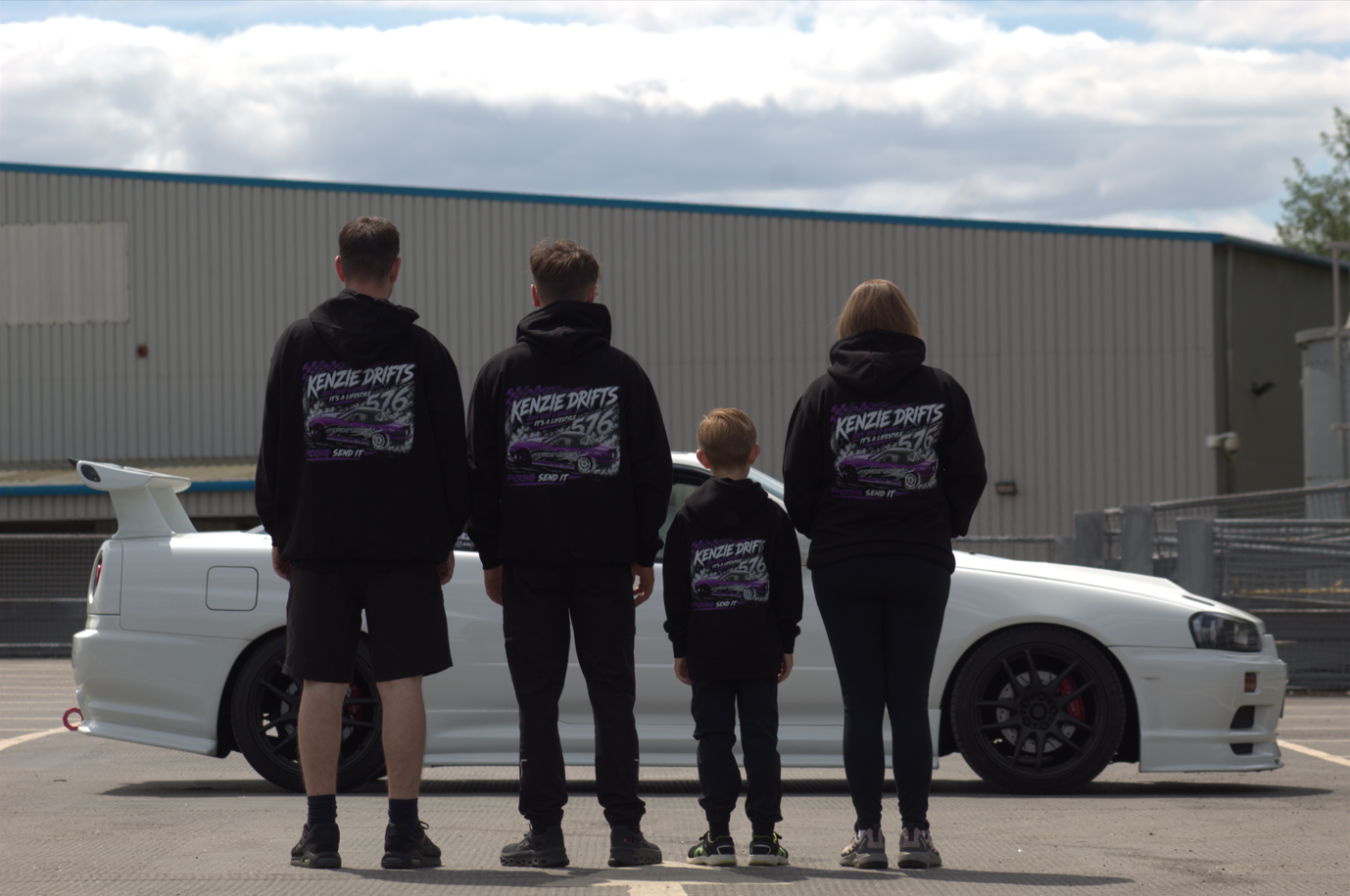 Family of four wearing matching black hoodies with purple graphics stands in front of a white sports car in a parking lot.