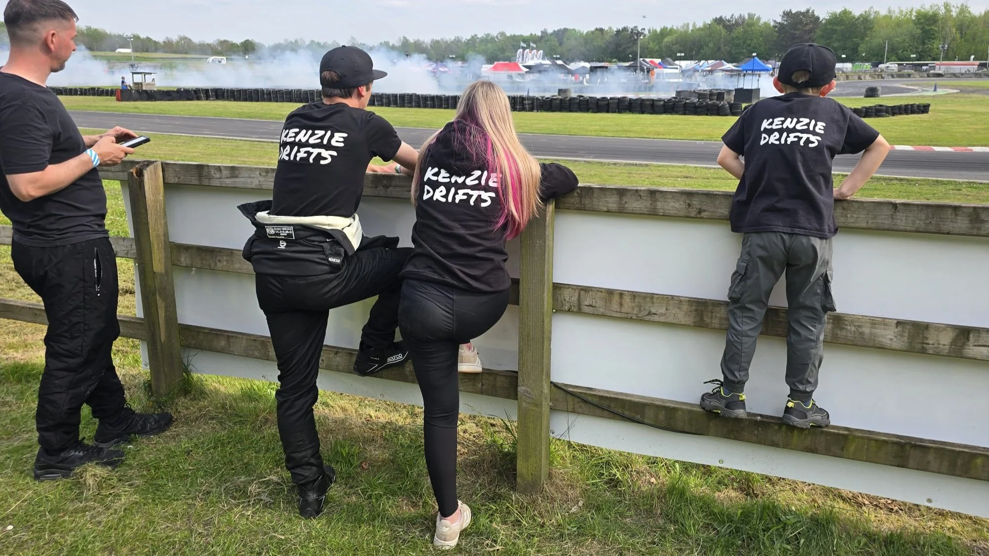 Four people watching an outdoor drift racing event from behind a wooden fence. Two are wearing black shirts with 'Kenzie Drifts' written on the back. Smoke and tire marks are visible on the racetrack in the background.