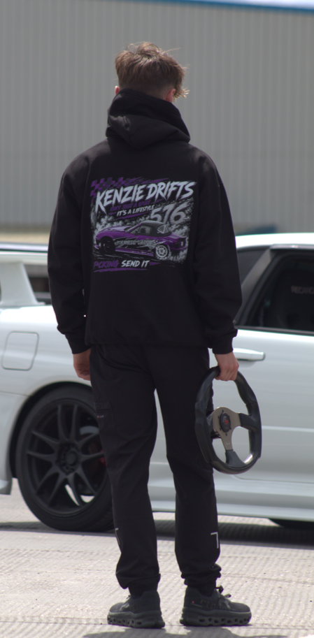 Person wearing a black hoodie holding a steering wheel standing in front of white cars and a metal building.
