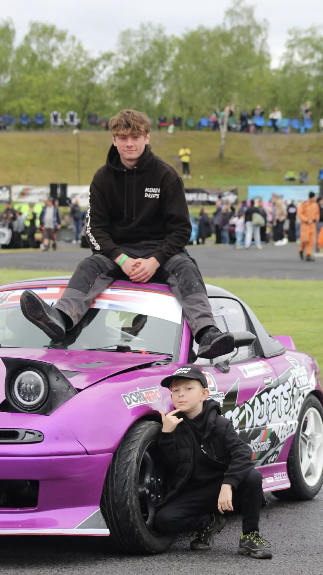 Two young boys at a motorsport event with a custom purple car decorated with sponsorship logos. One boy is sitting on top of the car, and the other is crouching next to the front tire, making a hand gesture.