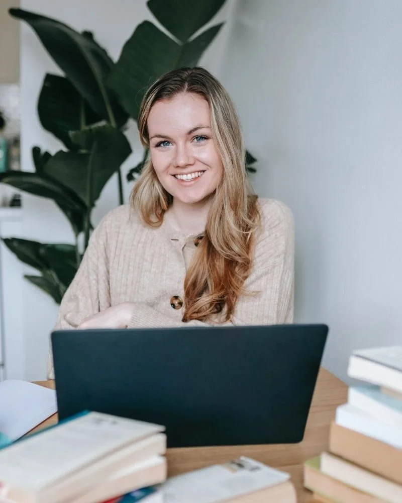 A young woman with long blonde hair and blue eyes smiling at her laptop surrounded by books on a desk, with a large green plant in the background.
