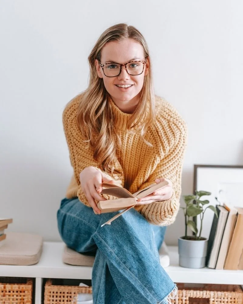 A woman with long blonde hair, glasses, wearing a yellow sweater and blue jeans, sitting on a white bench with a book in her hands in a cozy indoor setting.