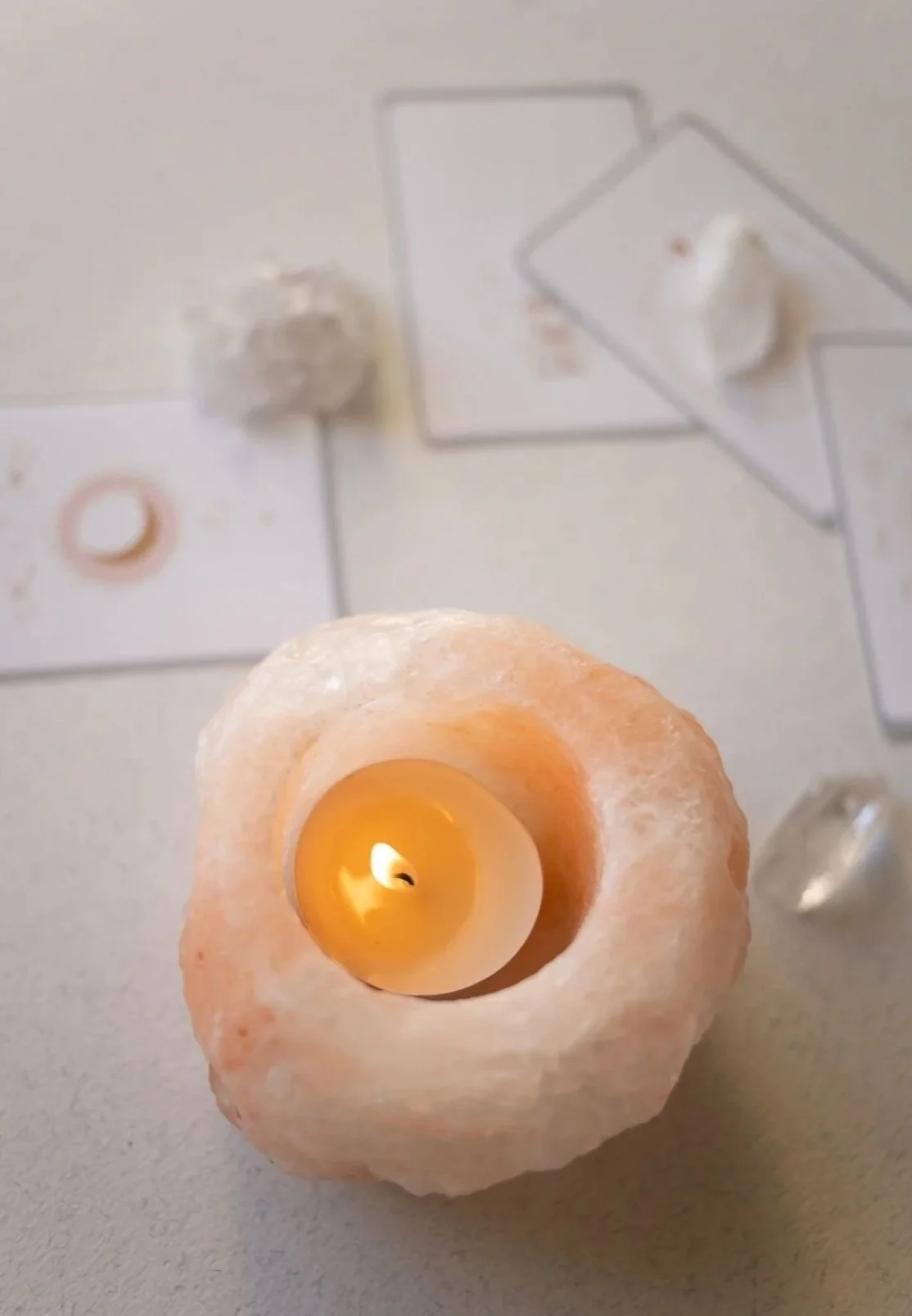 A lit candle inside a salt lamp holder on a table with scattered appearance, blurry papers with drawings or text, and small objects around.
