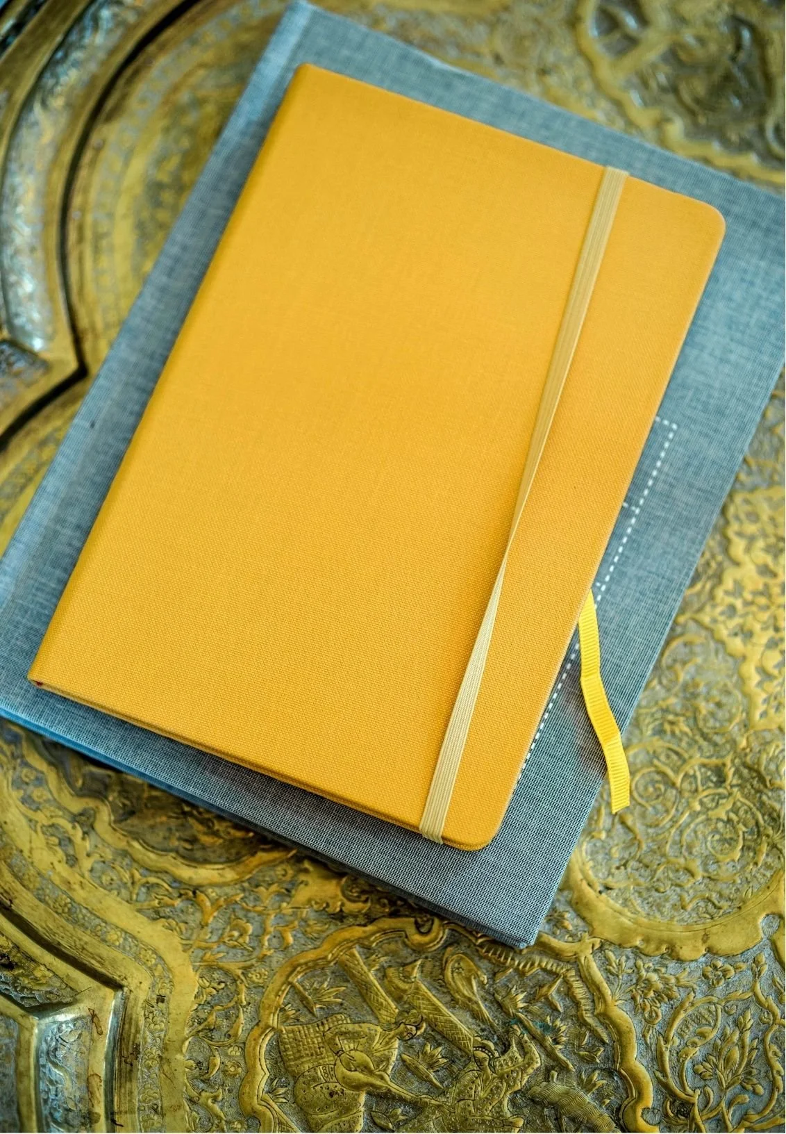 A yellow notebook with an elastic band and ribbon bookmark placed on top of a closed blue book, resting on a decorative gold and ornate table surface.