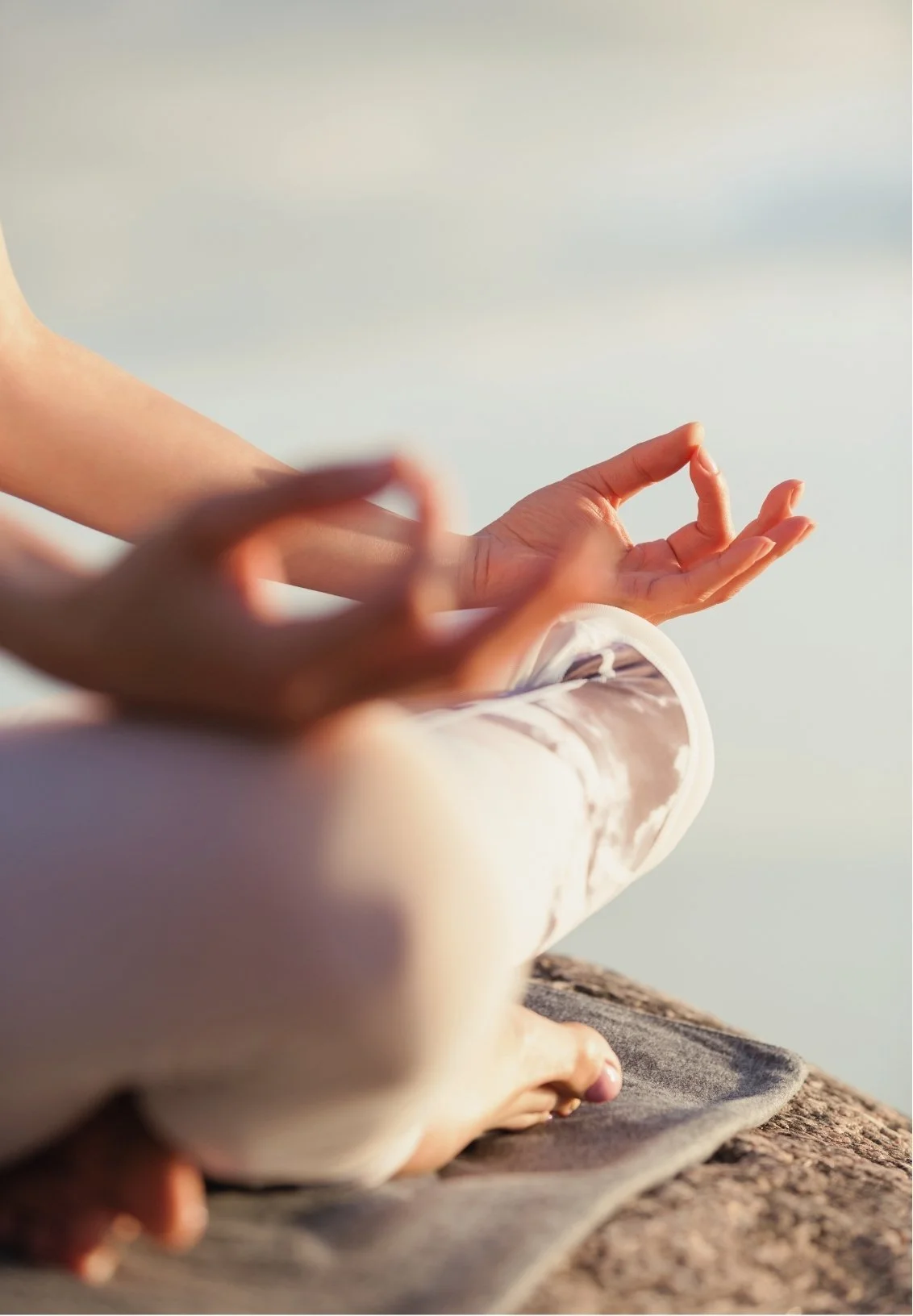 Person practicing yoga or meditation outdoors, sitting cross-legged with hands in a mudra gesture, on a rocky surface with a blurred background.