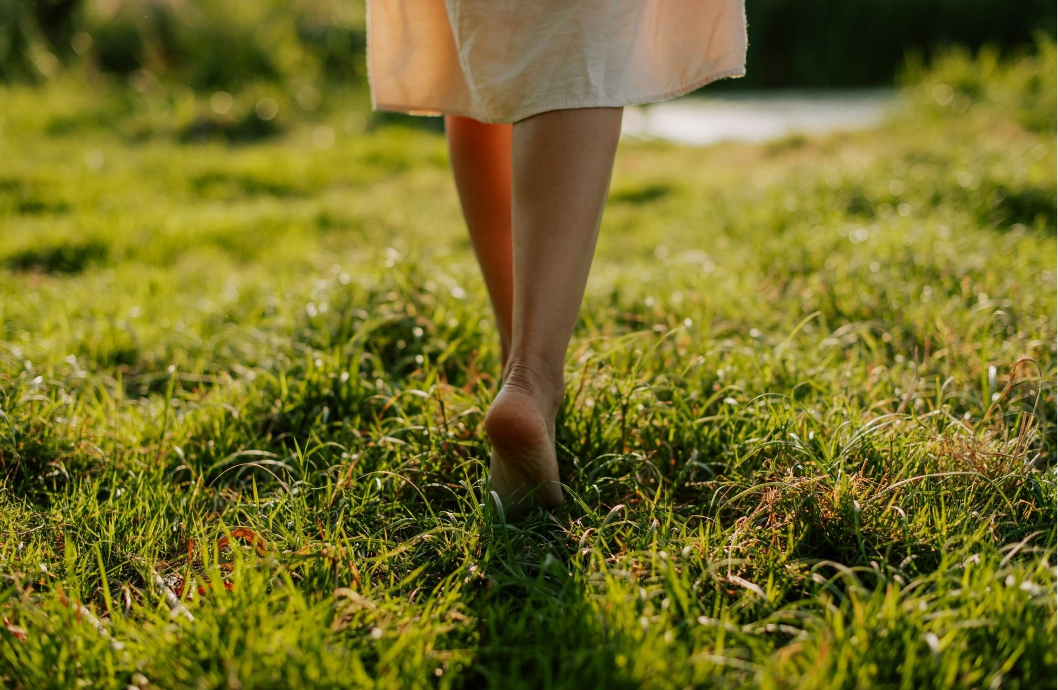 A person walking barefoot through a grassy field in sunlight, wearing a light-colored dress.