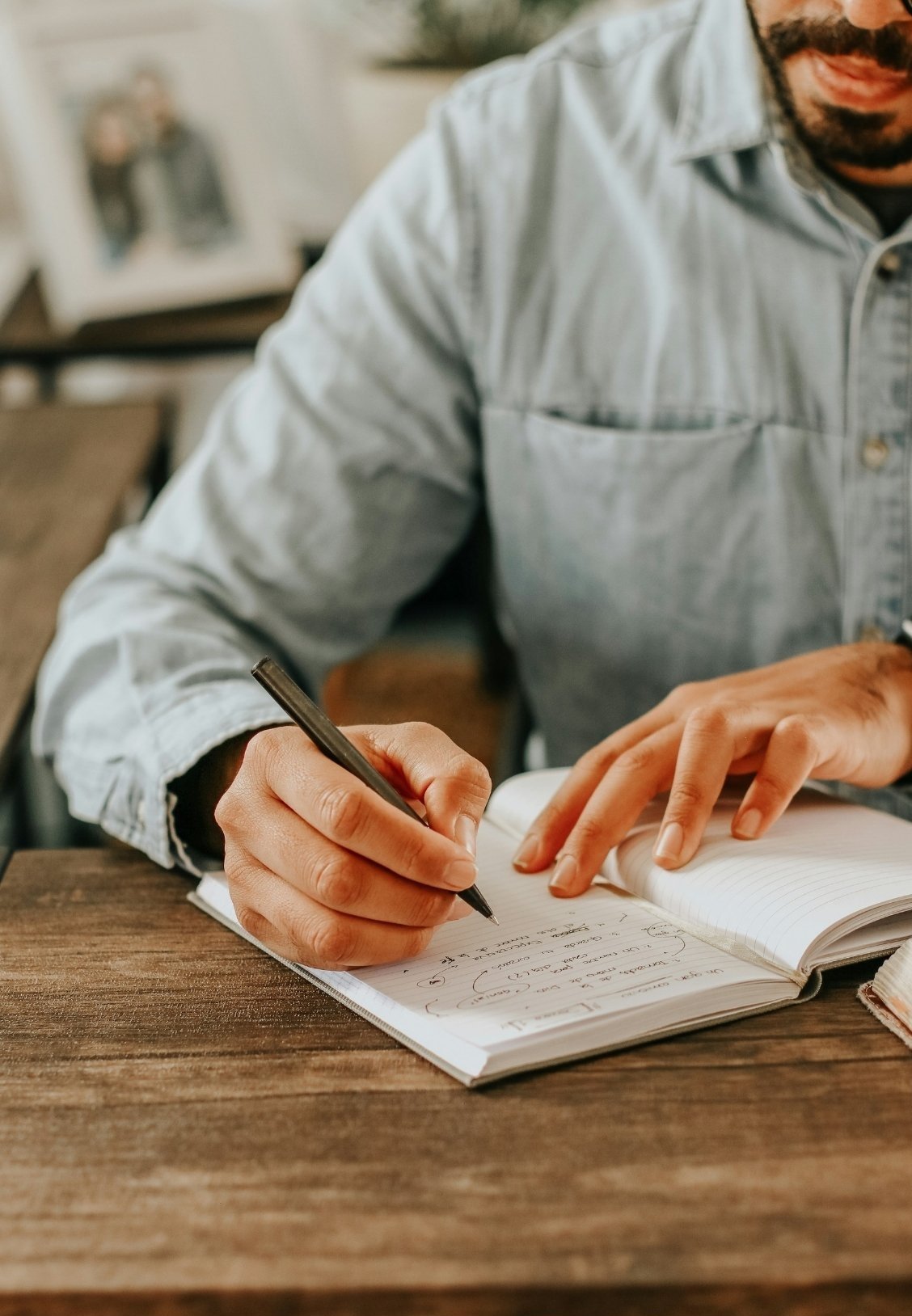 A person wearing a light denim shirt is writing in a notebook with a black pen. The focus is on the hand and notebook, with a blurred background of framed photos.