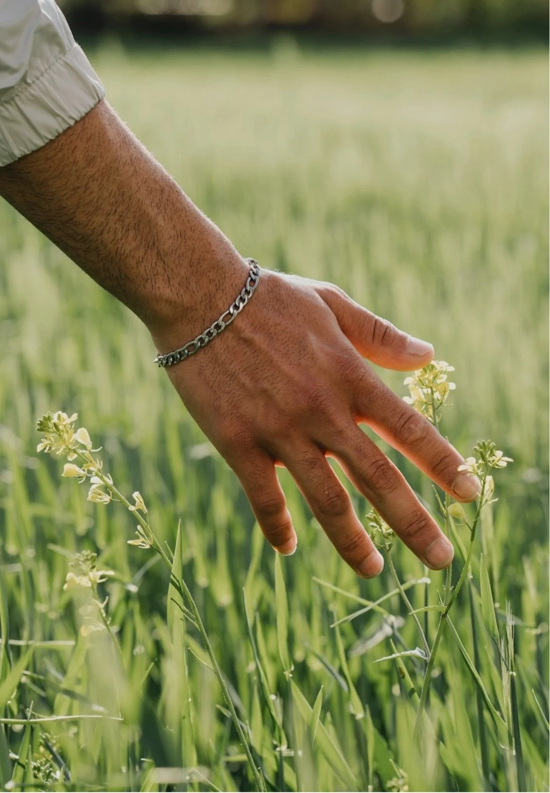 Close-up of a person's hand touching green grass and small white flowers in a field, wearing a silver chain bracelet.