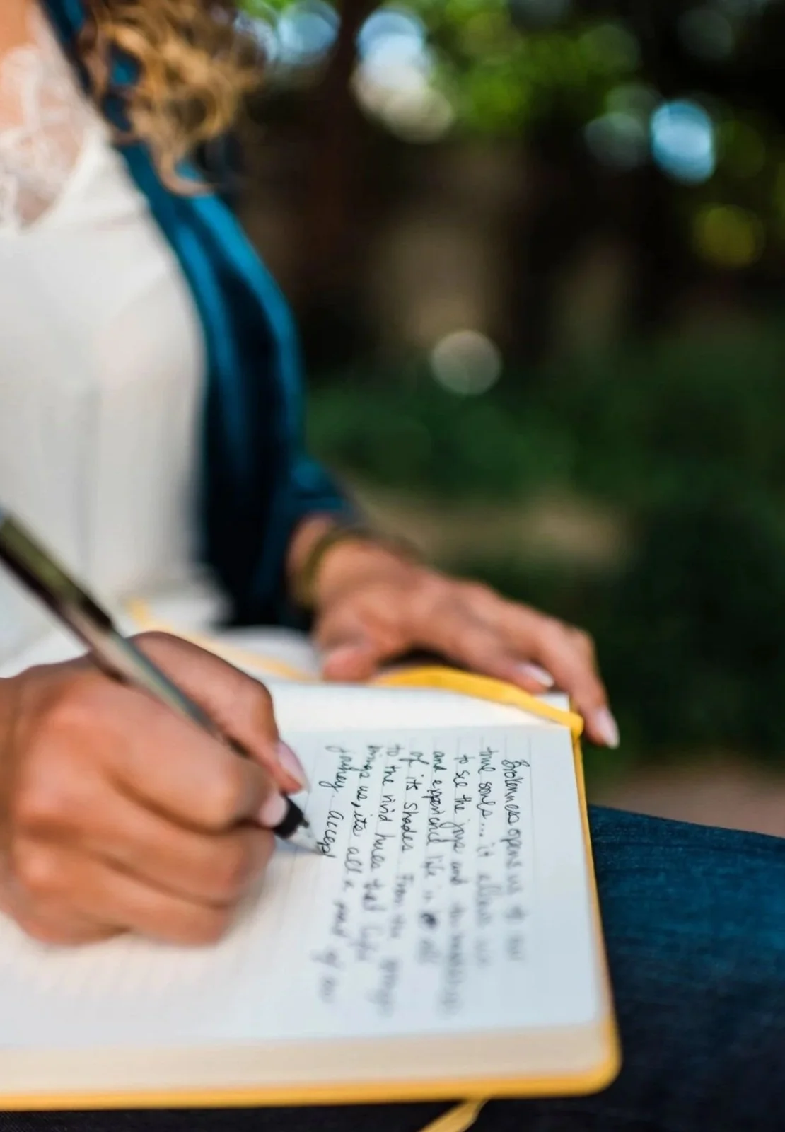 A person writing in a notebook outdoors, with a blurred background of trees and greenery.