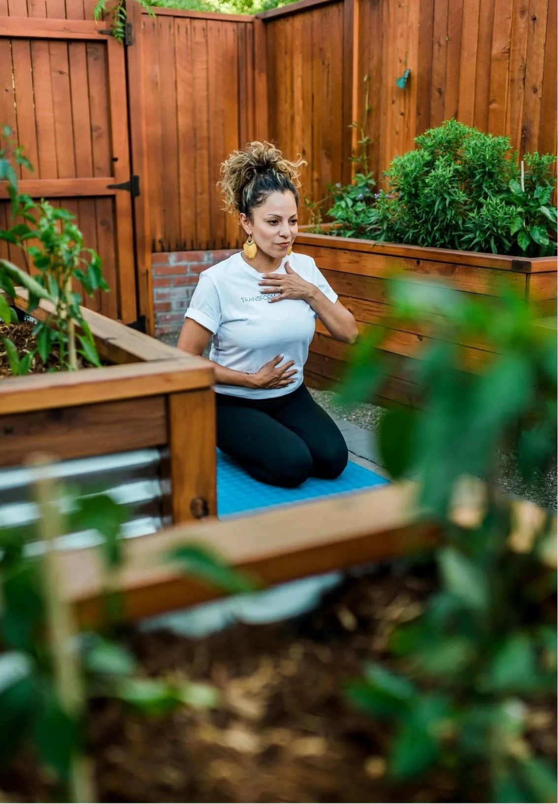 Mery Vargas Mares, therapist in Reno, NV, practicing yoga in a backyard garden, kneeling on a yoga mat with one hand on her chest and the other on her stomach, surrounded by wooden planters with green plants and a wooden fence.