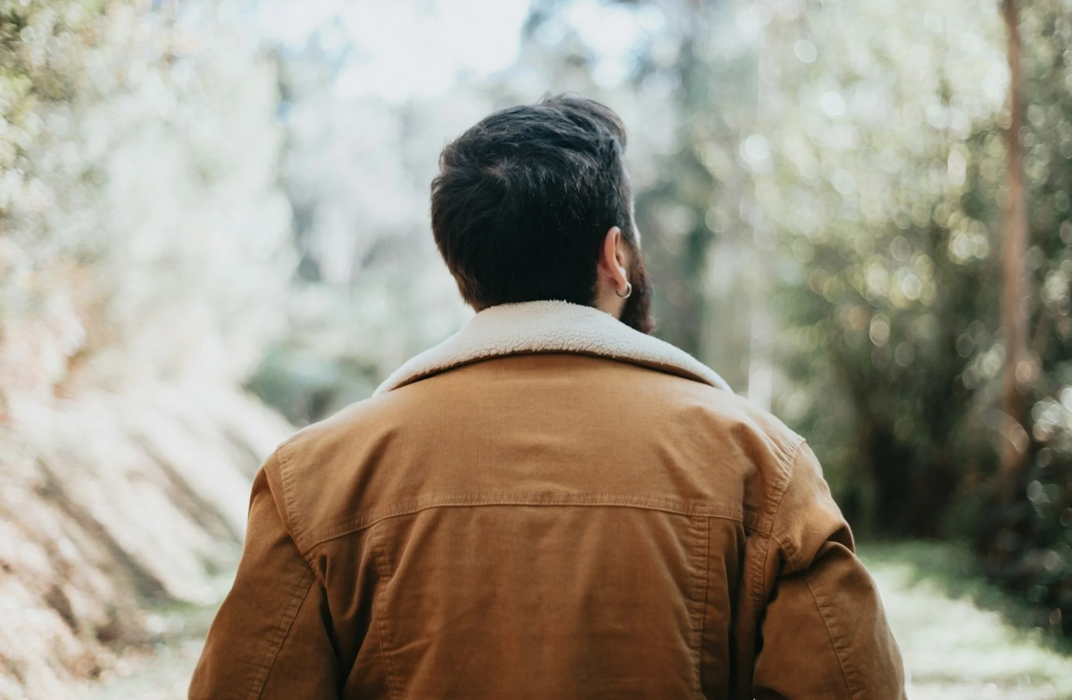 Rear view of a person with short dark hair and earrings, wearing a beige jacket with a white collar, standing outdoors in a forested area with trees and soft sunlight.