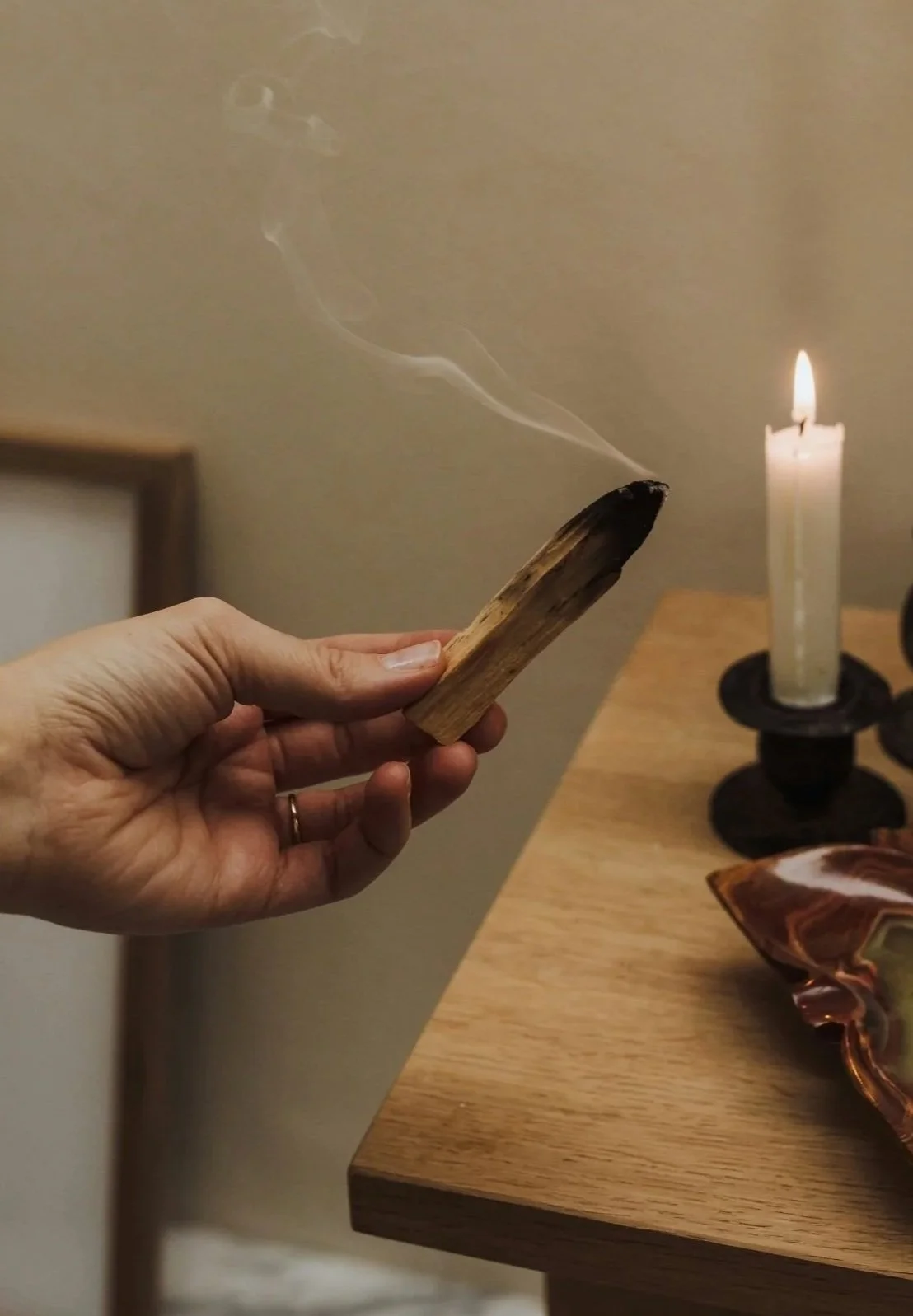 A hand holding a burning stick with smoke rising, in front of a lit candle on a wooden table.