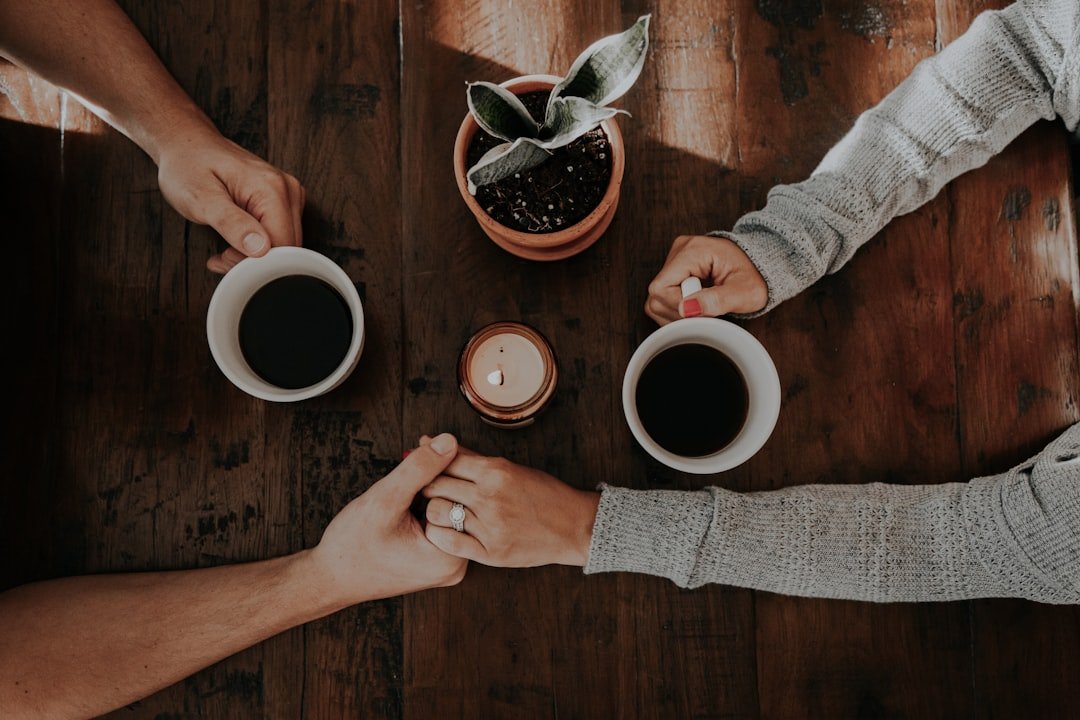 Two people holding hands across a wooden table, each with a cup of coffee, a potted plant, and a small lit candle between them.