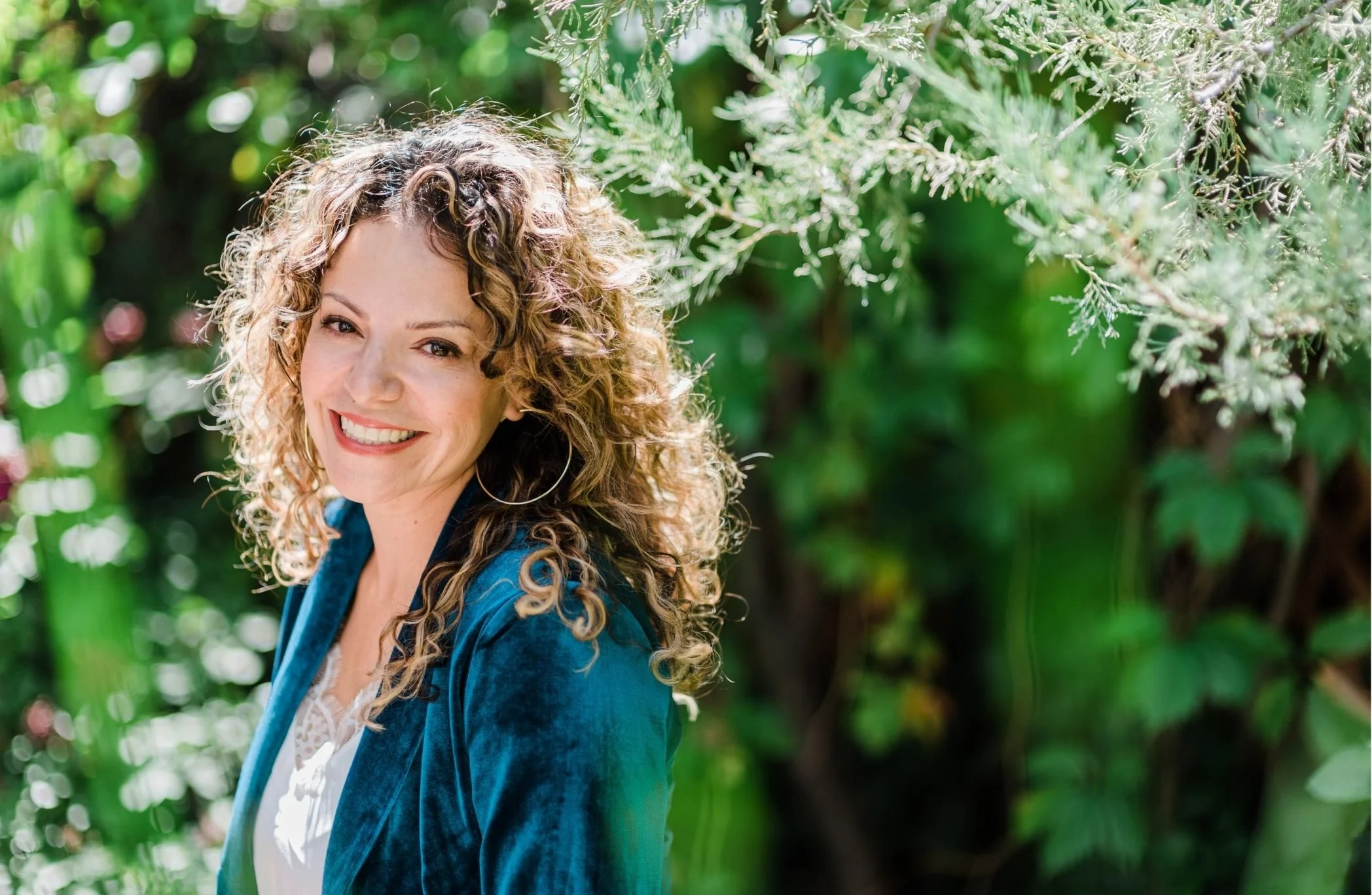 A woman with curly blond hair smiling in a lush green outdoor setting.