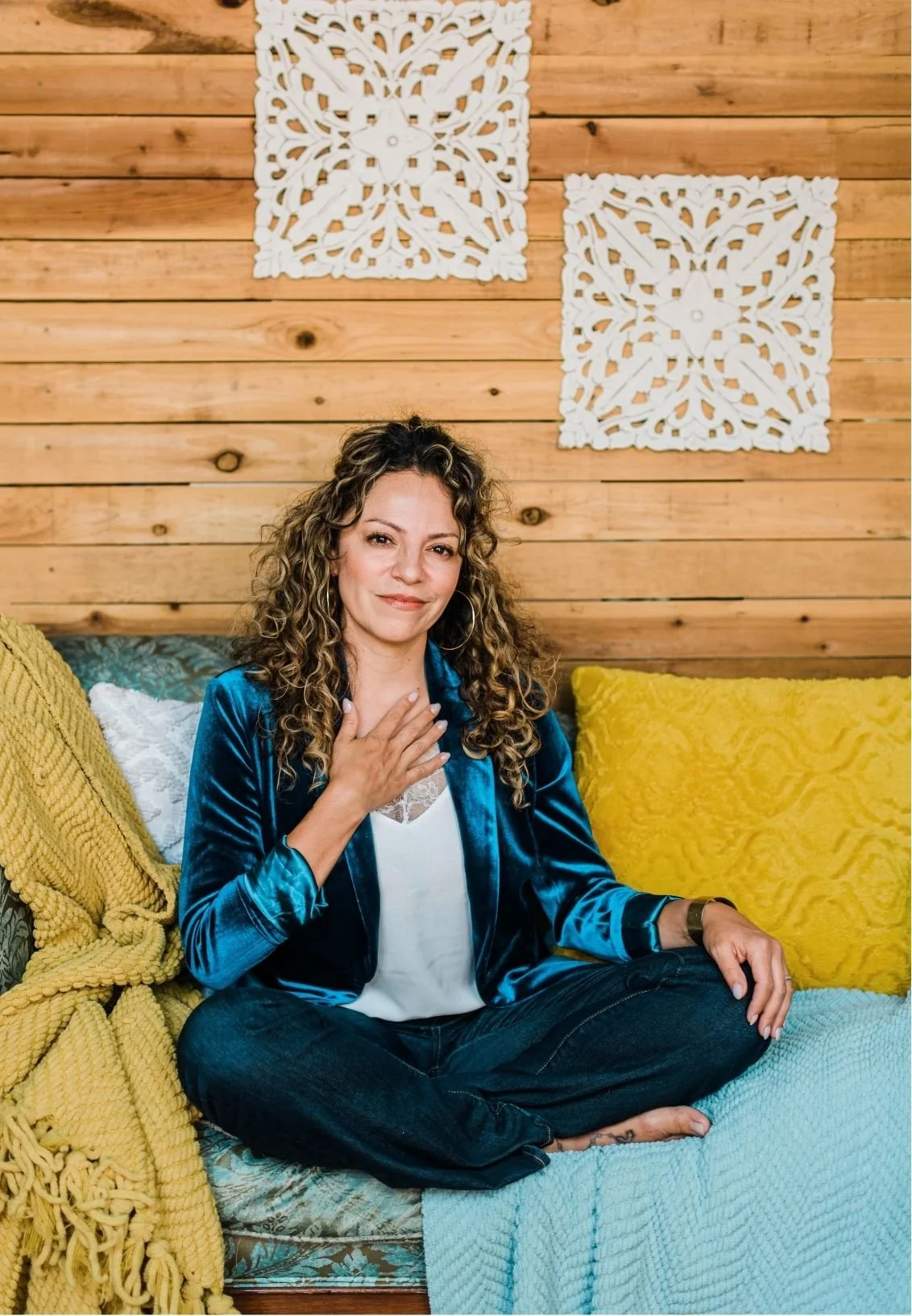 Mery Vargas Mares, therapist in Reno, sitting cross-legged on a couch in a cozy room with wooden walls and decorative white wall art. She is wearing a blue velvet jacket, a white top, and has curly hair. She is touching her chest with her left hand.