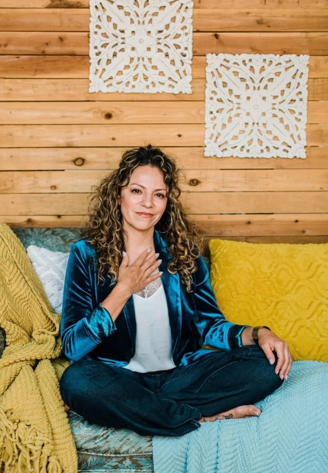 A woman with curly hair, wearing a blue velvet jacket and black pants, sitting cross-legged on a couch with yellow and white pillows. She has one hand on her chest and is smiling softly. The background features wooden paneling with two white decorative wall hangings.