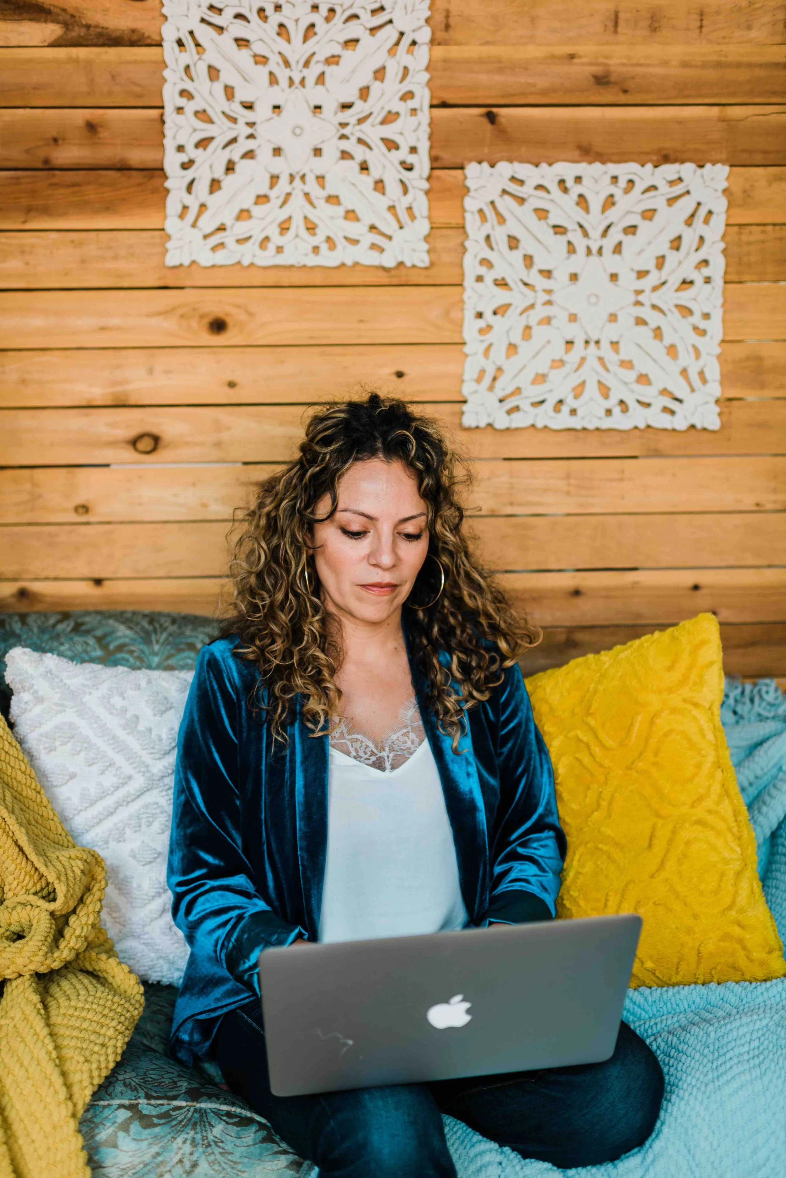 Mery Vargas Mares, therapist in Reno, NV, with curly hair and earrings sitting on a sofa in front of a laptop, surrounded by colorful pillows, with a wooden wall and decorative white panels in the background.
