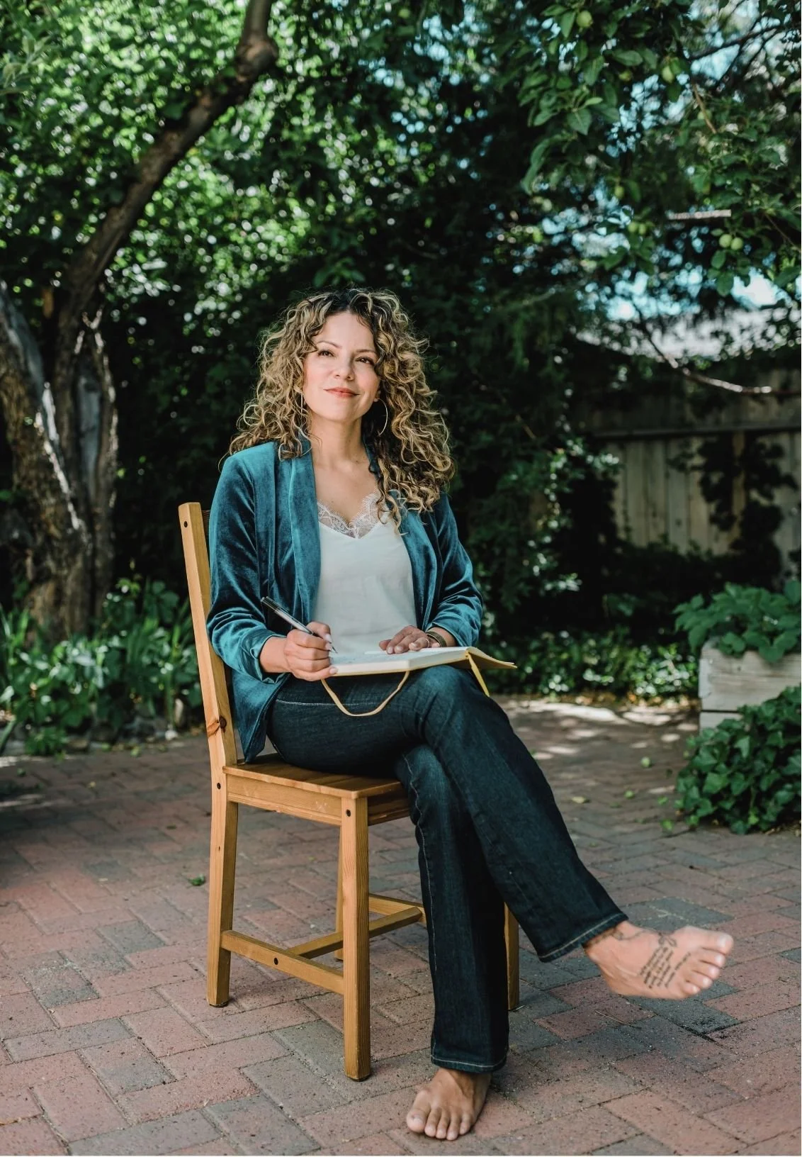 A woman with curly hair sitting outdoors on a wooden chair, holding a notebook and a pen, surrounded by greenery and trees.