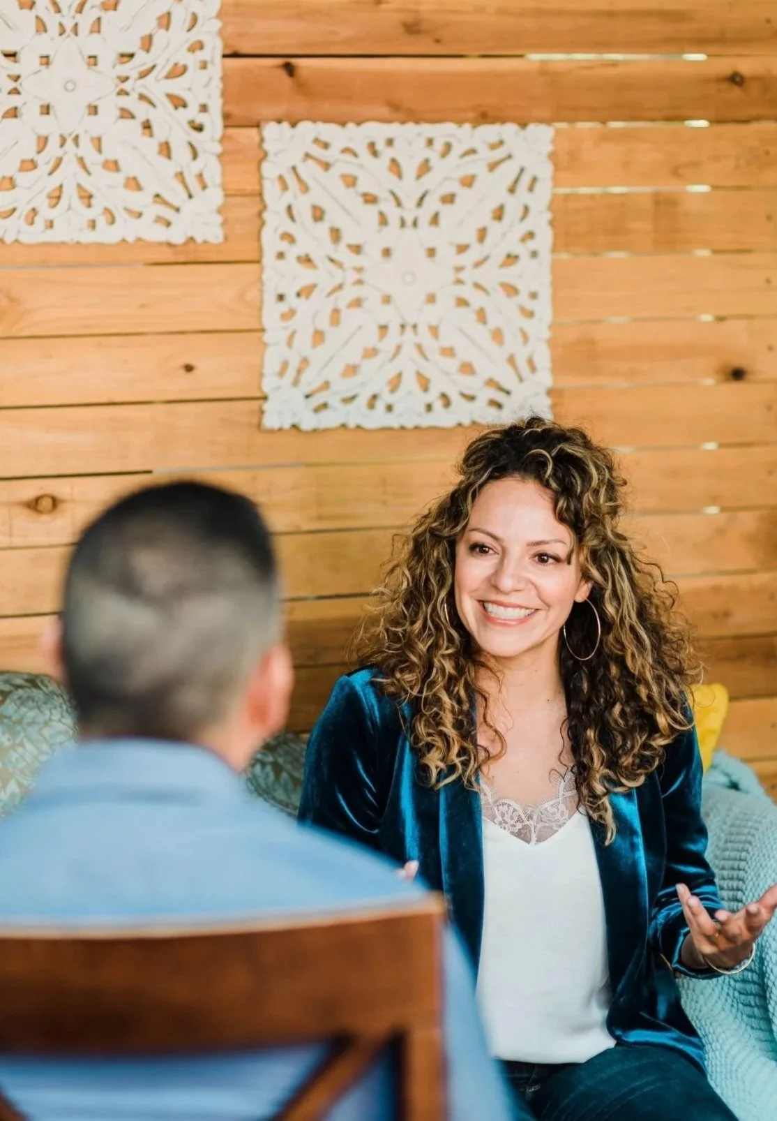 A woman with curly hair, wearing hoop earrings and a blue jacket, smiling and talking to a man with short hair, sitting indoors with a wood-paneled wall and white decorative wall hangings.